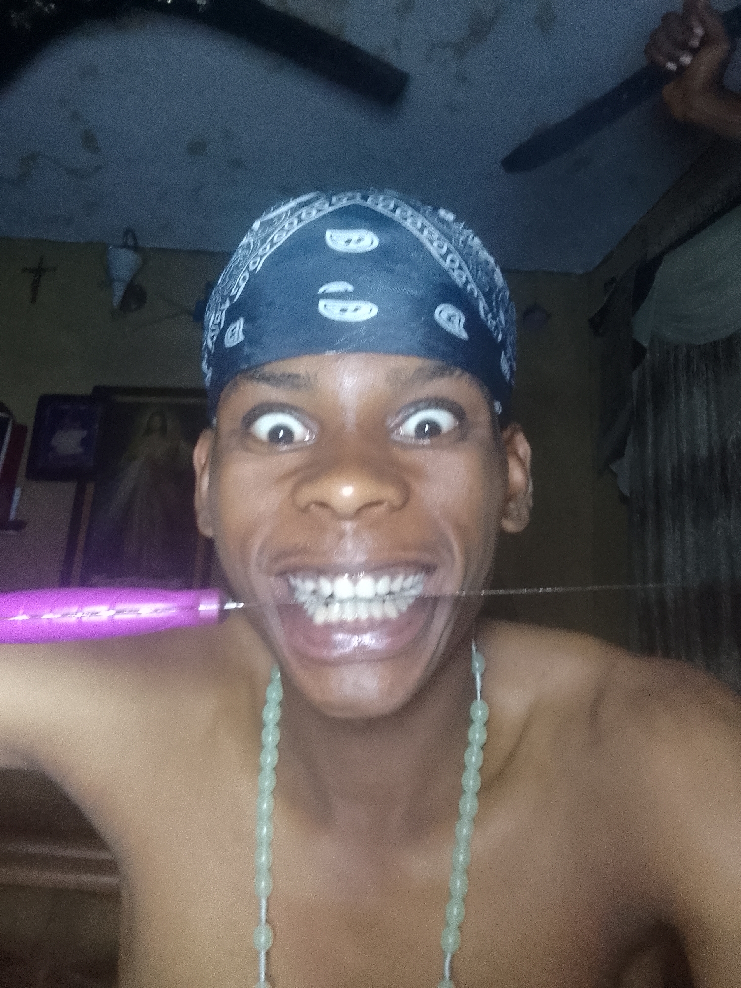A close-up shot depicts an individual wearing a blue bandana and a beaded necklace, holding a purple object in their mouth. The person has wide eyes and an exaggerated smile, exposing their teeth. The background includes a cross hanging on the wall, framed pictures, and a ceiling fan. The location is Nkpor, Nigeria.
