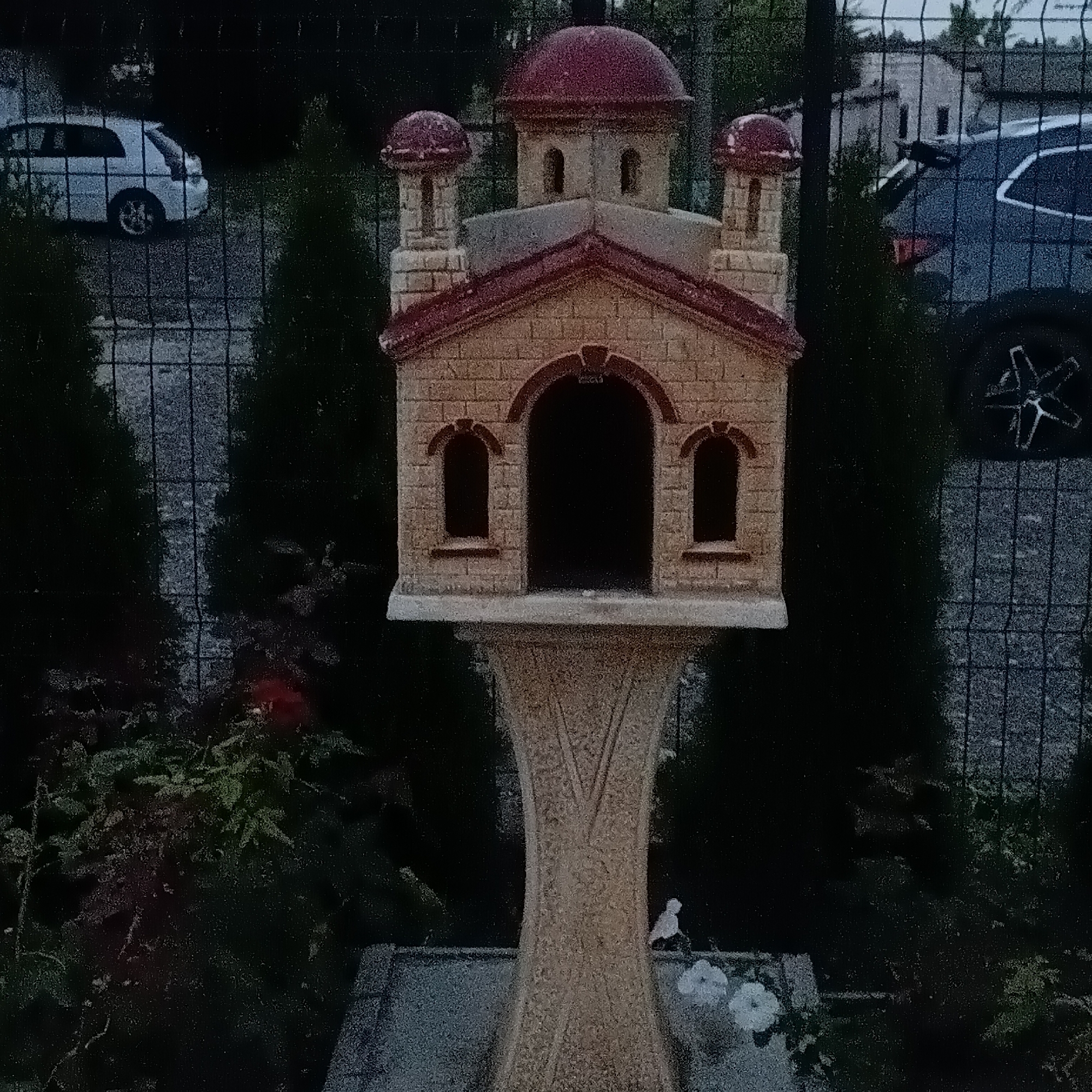 This image depicts an outdoor scene, likely in a residential area or garden, in the city of Chișinău, Moldova. The foreground is dominated by a decorative, multi-tiered structure resembling a miniature church or a birdhouse, constructed from what appears to be stone or concrete, with a red, domed roof. This structure stands on a pedestal, also made of similar material, exhibiting carved decorative patterns. In the lower part of the image, there are hints of foliage, possibly a garden, with some white flowers visible.
Behind the decorative structure, a wire fence runs horizontally, obscuring the background to some extent. Through the fence, several elements are visible. To the left, a white van or minivan is parked on a paved surface. Further back and to the right, a dark-colored SUV with its rear hatch open is parked. The background also shows glimpses of buildings and trees, suggesting an urban or suburban environment. The lighting suggests it is either early morning or late evening, with low ambient light conditions. No people are visible in the scene. The overall impression is one of a quiet, possibly residential setting with decorative elements.