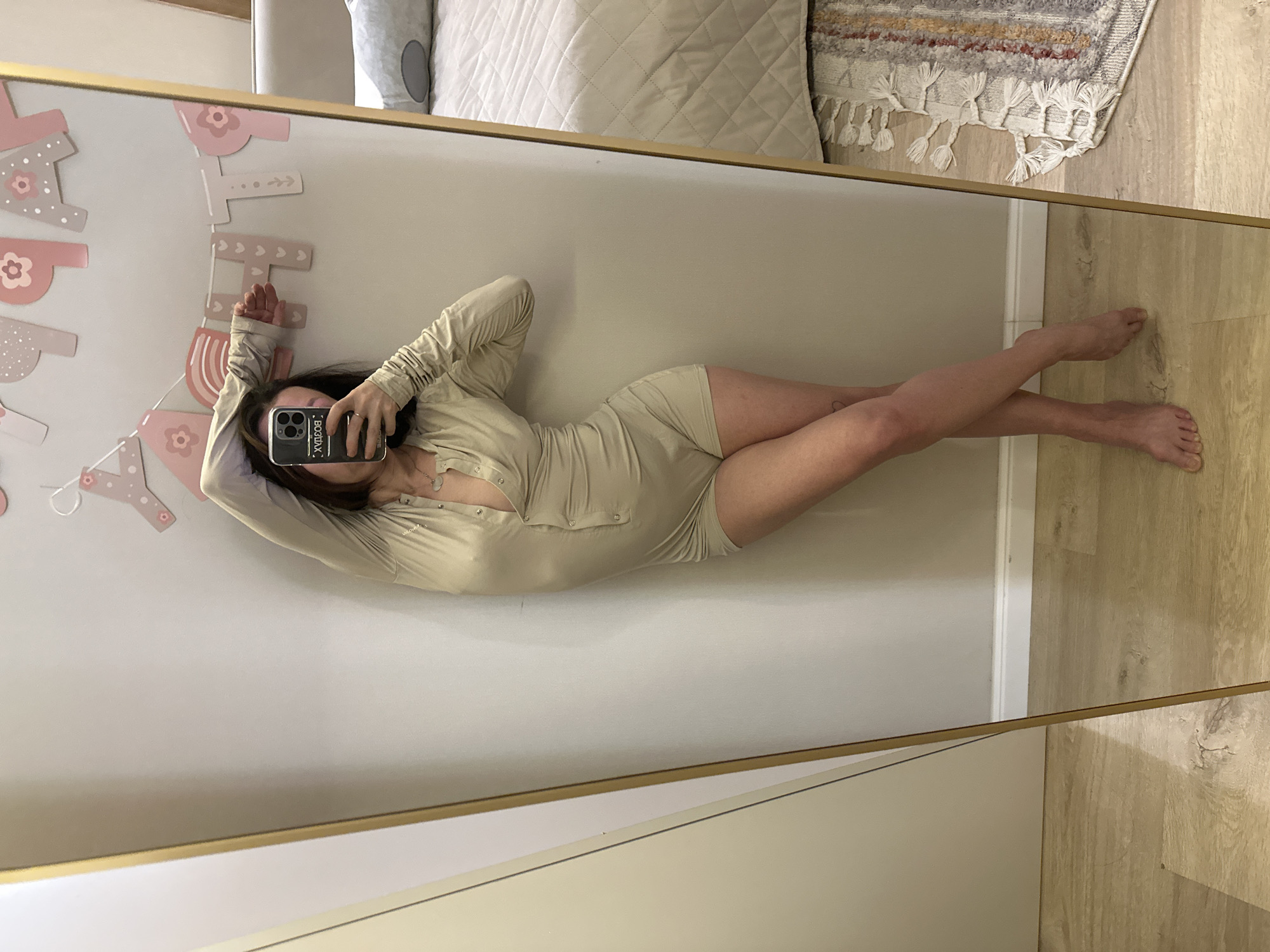 A woman is reflected in a mirror, positioned horizontally and wearing a light beige, long-sleeved romper with button detailing down the front. She is holding a mobile phone with a black case, obscuring her face in the reflection. Her hair is dark, and she wears a necklace. She is lying on her side with her legs crossed.
The mirror is framed in gold. In the background of the reflection, parts of a bed with a white quilted comforter, a throw rug, and a pink and white banner are visible. The floor appears to be light wood-toned.
There is no evidence of the location being Votkinsk, Russia.