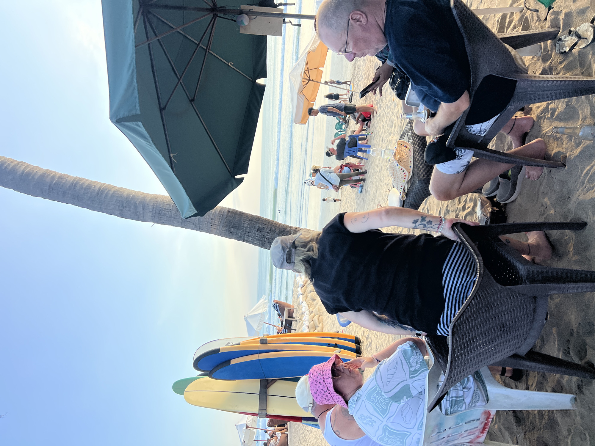 The image depicts a beach scene in Kuta, Indonesia, during what appears to be the late afternoon or early evening. Several individuals are seated at tables on the sand, with the ocean and waves visible in the background.
In the foreground, three people are seated at a table. A person wearing a pink bucket hat and sunglasses is on the left, facing the table. To their right, another person is seated with their back to the camera, wearing a black t-shirt and striped shorts. A third person is seated on the right, facing left and looking at a phone, wearing a dark blue shirt and glasses. Other people are visible further in the background, some interacting and others relaxing.
Objects in the scene include chairs, tables, surfboards propped up, umbrellas providing shade, bottles, and a straw hat. The environment is sandy, with a clear sky and the ocean forming the backdrop. The overall impression is one of a relaxed beachside gathering.