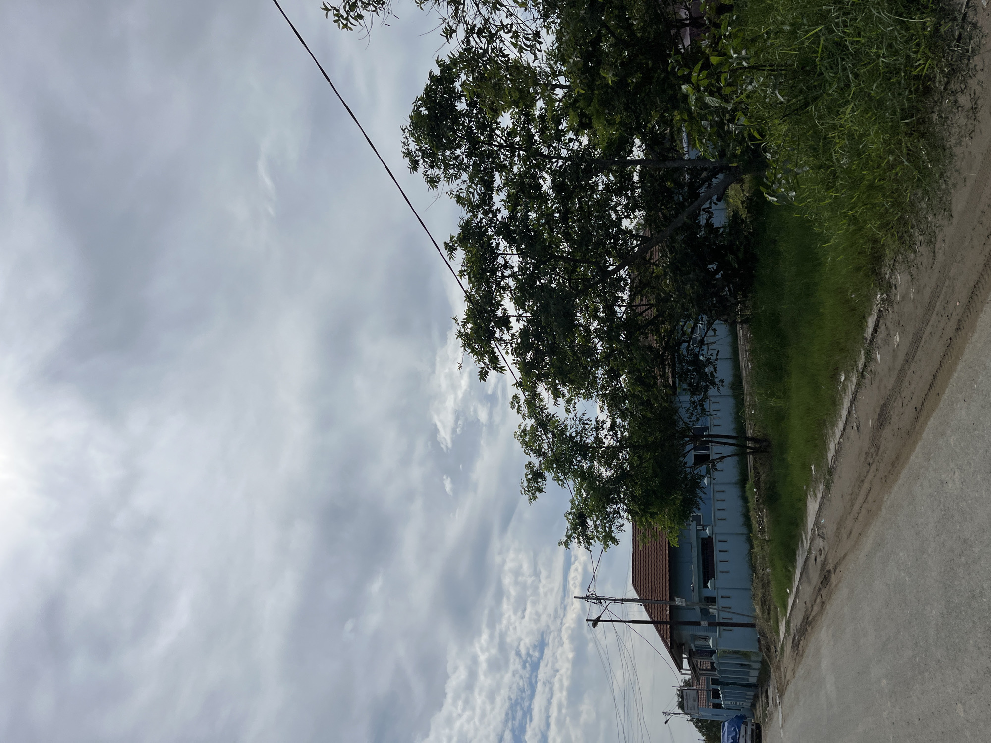 The image captures a cloudy day in Kota Kuala Simpang, Indonesia. A light blue building with a reddish-brown tiled roof is visible on the left, bordered by a light blue fence. In front of the building, there is a patch of green grass. Several trees with dense green foliage are positioned in front of the building and to the right, obscuring parts of the structure. A dirt and asphalt road runs along the bottom of the frame, with tire tracks visible on the dirt portion. Overhead, power lines stretch across the sky. The sky is filled with a mix of white and gray clouds, with some patches of bright sky suggesting sunlight filtering through. There are no people or animals visible in the scene.
