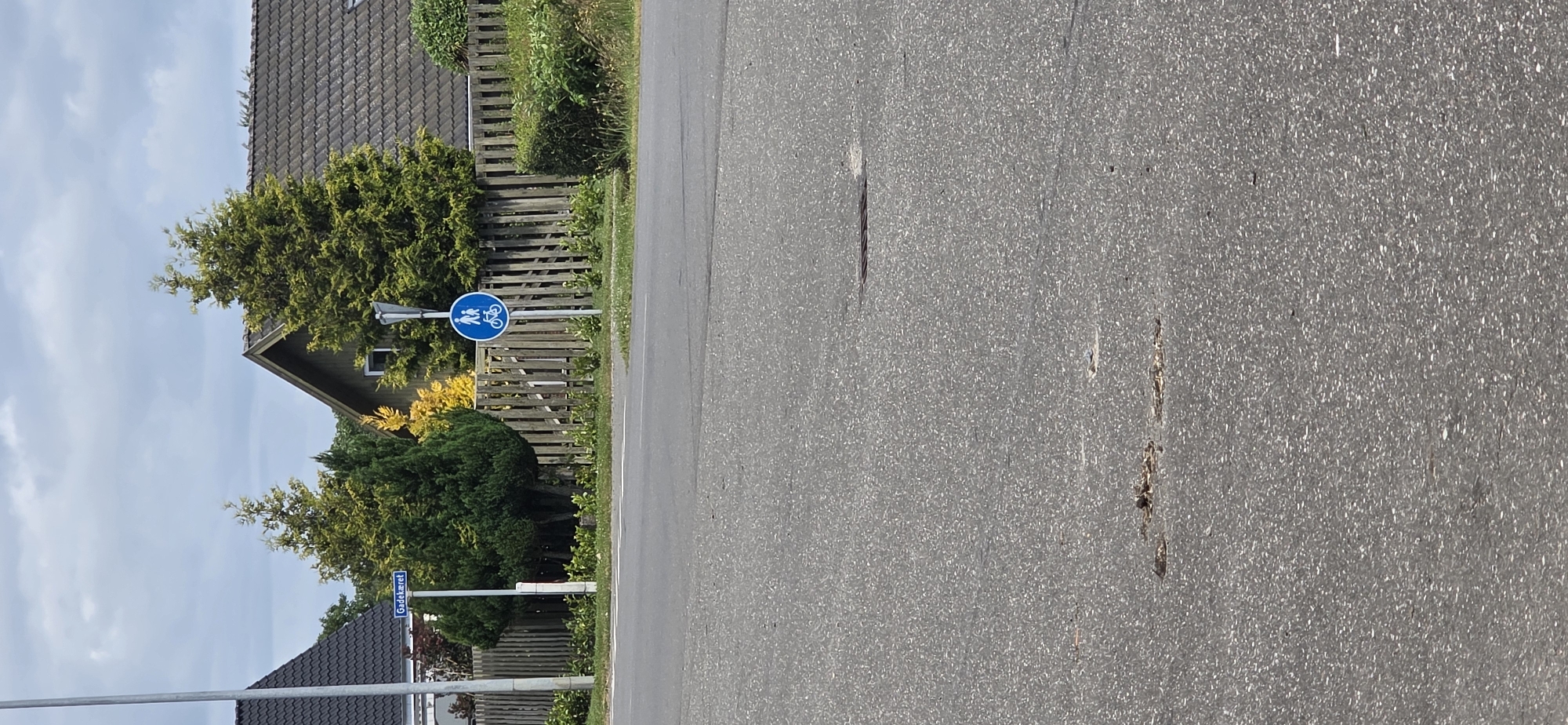 This is a photograph of a street scene in Hjørring, Denmark. In the foreground, a paved road with some debris and a manhole cover is visible. In the mid-ground, there is a sidewalk or curb with grass and some small plants. A wooden fence runs along the edge of the property. Behind the fence, there are trees with green and yellow foliage, and the roofs of houses. A street sign that reads "Gadekjæret" is mounted on a pole. Next to it, a blue circular sign with white silhouettes of a pedestrian and a cyclist indicates a shared path. The sky is overcast with scattered clouds.
