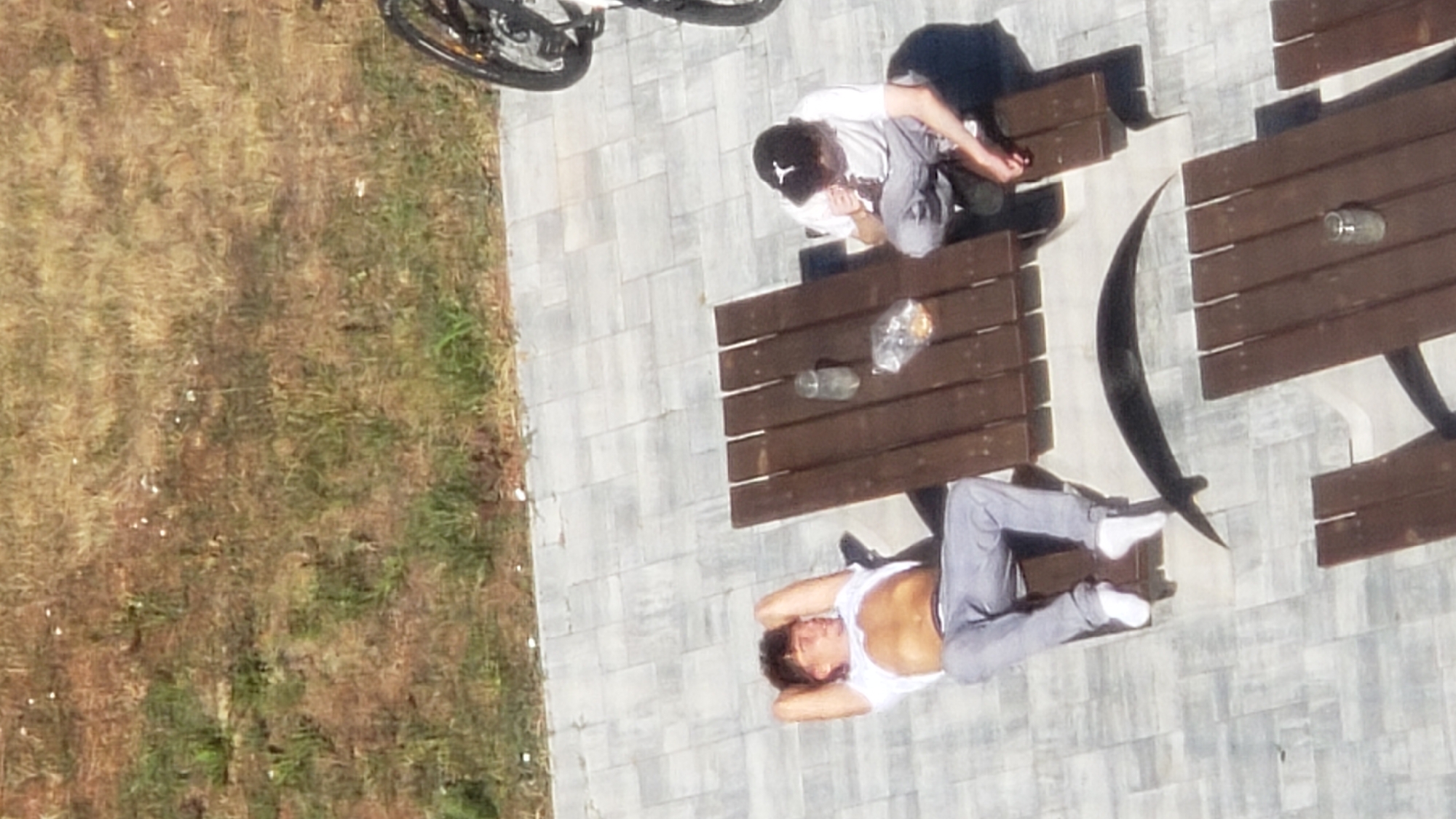 Two young men are seen outdoors on a paved area near a grassy slope. One man, lying down with his hands behind his head and a white t-shirt exposing his midriff, is on a chair. The second man, wearing a white t-shirt and a black baseball cap, is seated at a wooden picnic table. A bag of snacks, a glass jar, and a bicycle are also visible in the scene. The image is taken from a high angle, suggesting it was captured from a building or elevated position. The setting appears to be a recreational or public space within the city of Prešov, Slovakia, likely a park or outdoor seating area.