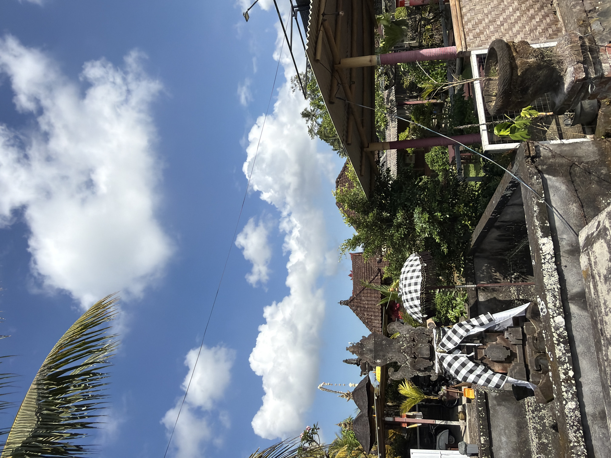 This is a photograph of a traditional Balinese outdoor setting, likely a temple complex or a sacred compound. The scene is dominated by a bright blue sky with scattered white, puffy clouds, suggesting a sunny day, possibly mid-morning or early afternoon given the lighting. Lush green foliage and trees are visible, interspersed with traditional Balinese architecture featuring tiled roofs and bamboo structures.
The foreground features a stone structure, possibly an altar or a decorative element, adorned with black and white checkered fabric, known as *poleng*. This fabric is a significant cultural symbol in Bali, often used in religious ceremonies and to represent the balance of opposing forces. A small umbrella, also in the *poleng* pattern, is displayed, further emphasizing the cultural context. In the lower left corner, a frond of a palm tree is visible, adding to the tropical atmosphere.
In the background, more buildings with intricate roof designs can be seen, along with more greenery. The overall impression is one of a serene and culturally rich environment. There are no visible people in the frame, suggesting the photograph was taken at a moment of quiet observation. The location cues, particularly the *poleng* fabric and the architectural style, strongly indicate a setting in Bali, Indonesia, consistent with Tampaksiring, which is known for its temples and cultural heritage.