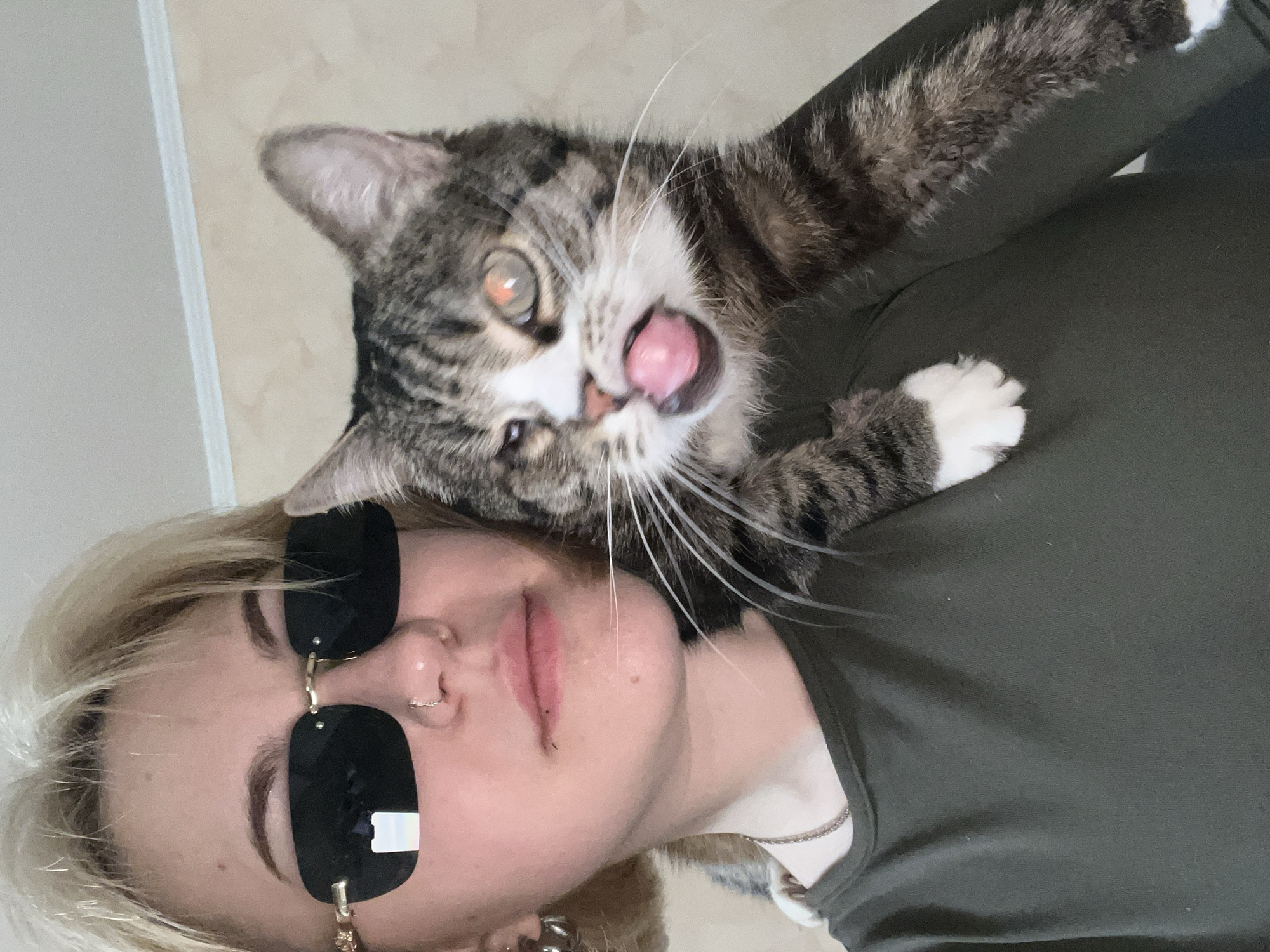 A selfie shows a fair-skinned person wearing sunglasses and a green shirt, with a tabby cat draped over their shoulder. The cat is licking its lips and looks towards the camera. The location appears to be indoors with a neutral-colored wall in the background. The city and specific address are unknown.