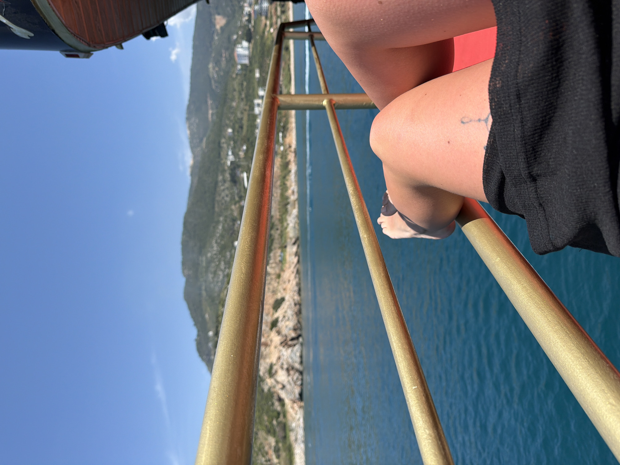 The image presents a view from a high vantage point, possibly a cable car or similar structure in the city of Alanya, Turkey. The perspective is angled downward, showcasing a landscape of mountains, coastline, and the blue waters of the Mediterranean Sea. A part of the city is also visible in the distance.
A golden safety railing is prominently featured in the foreground. Adjacent to it, a person's legs and feet are visible, with the individual wearing what appears to be a dark-colored cover-up or clothing. A small tattoo is discernible on the person's upper leg.