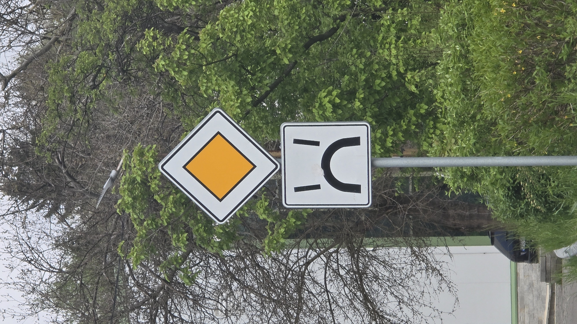 Two road signs are affixed to a metal pole. The sign on the left is diamond-shaped, white with a black border, and has an orange diamond in the center. The sign on the right is square, white with a black border, and depicts a road with a sharp U-turn to the right. The background consists of trees and foliage. A glimpse of a white building and dark car is visible at the bottom right. The location is Pernik, Bulgaria.
