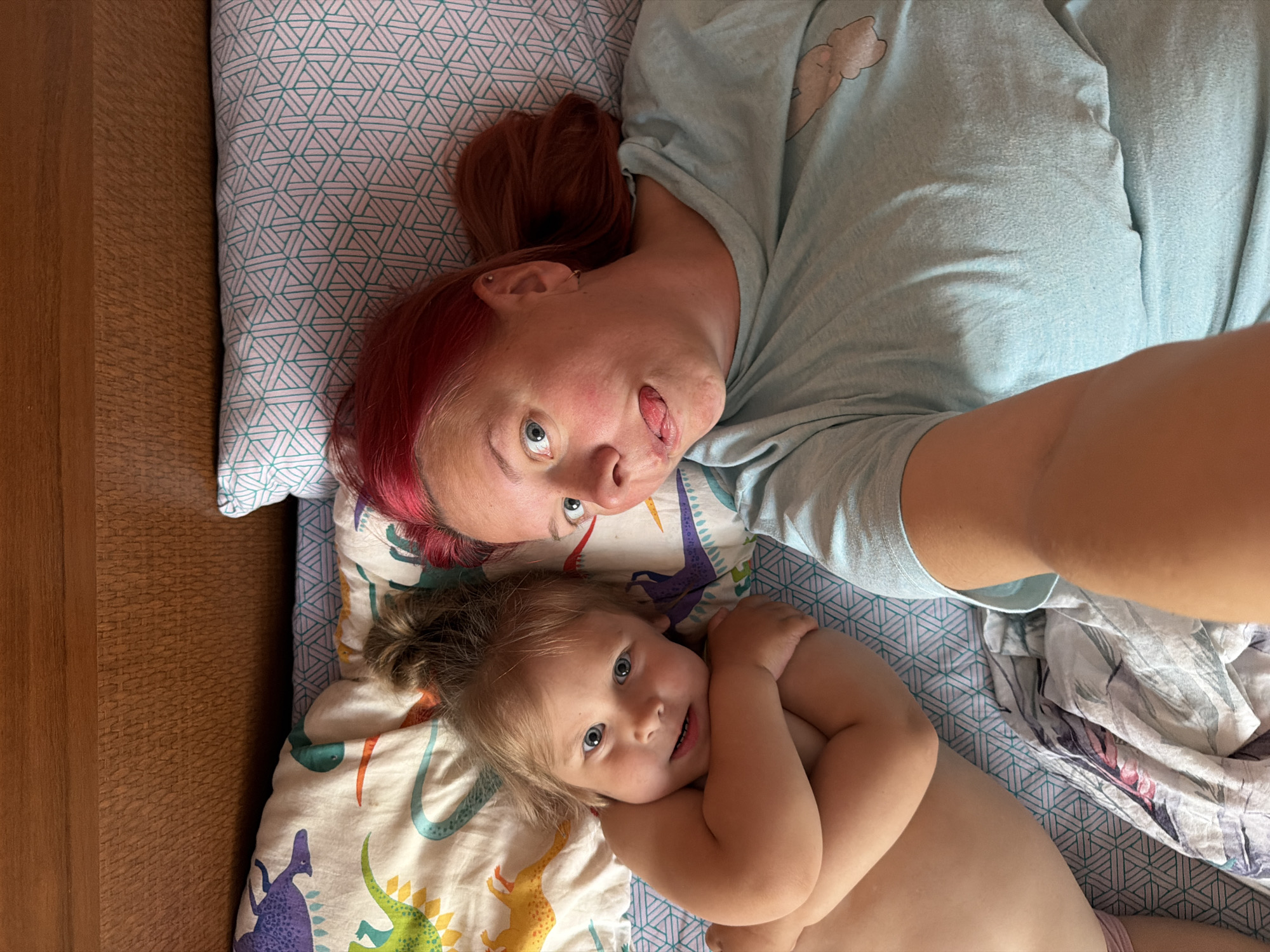 An overhead shot shows two people lying on a bed. On the left, a young child with blonde hair and blue eyes is positioned on their back, with their arms crossed over their chest. The child is looking directly at the camera with a slight smile. To the right of the child is an adult with bright red hair, also looking at the camera. The adult is sticking their tongue out and appears to be taking a selfie. Both individuals are lying on a bed with patterned sheets. The background features a wooden headboard. The scene is indoors and appears to be a bedroom.