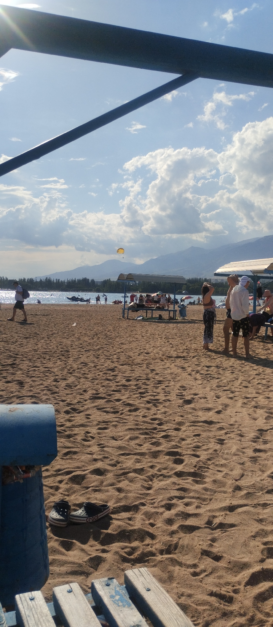 The image depicts a sunny day at a beach in Cholpon-Ata, Kyrgyzstan. The foreground shows a sandy beach with a blue trash bin on the left and a wooden bench in the lower part of the frame. A pair of black Crocs sandals are visible on the sand near the trash bin. In the mid-ground, several people are scattered across the beach and near the water. Some are standing, some are sitting, and a group is gathered under a shaded structure. In the water, there are several boats, including a jet ski. Further out, a yellow parasail is visible in the sky, being towed by an unseen boat. The background features a calm body of water, a tree line, and distant mountains under a cloudy sky with patches of blue. The overall scene is one of a relaxed beach day with recreational activities.