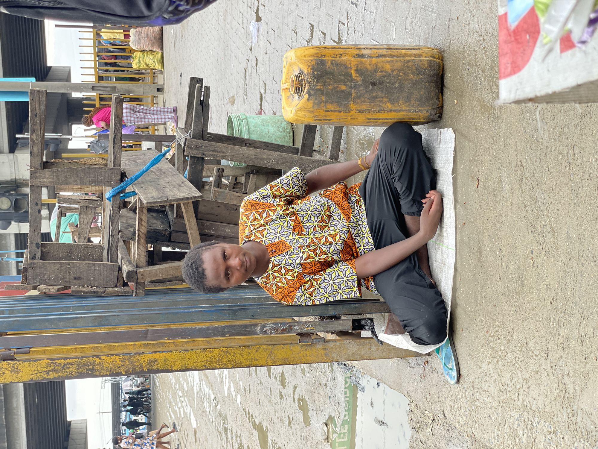 The image depicts a street scene in Lekki, Nigeria. In the foreground, a young person sits cross-legged on a white mat on a wet, concrete ground. They are wearing a short-sleeved shirt with an orange, white, and yellow geometric pattern and black pants. To the right of the person is a large, yellow plastic container. Behind the person and to their left, a tall, yellow and blue metal gate is partially visible, leaning against a structure made of rough wooden planks. In the background, an overhead bridge is visible, with traffic and people moving beneath it. The ground is uneven and appears to have puddles of water and debris, suggesting recent rain or poor drainage. Further in the background, there are more structures, yellow fencing, and various sacks, possibly indicating a market area. A person in a pink shirt and plaid pants is seen further in the background, standing near the fence. The lighting suggests an overcast day.