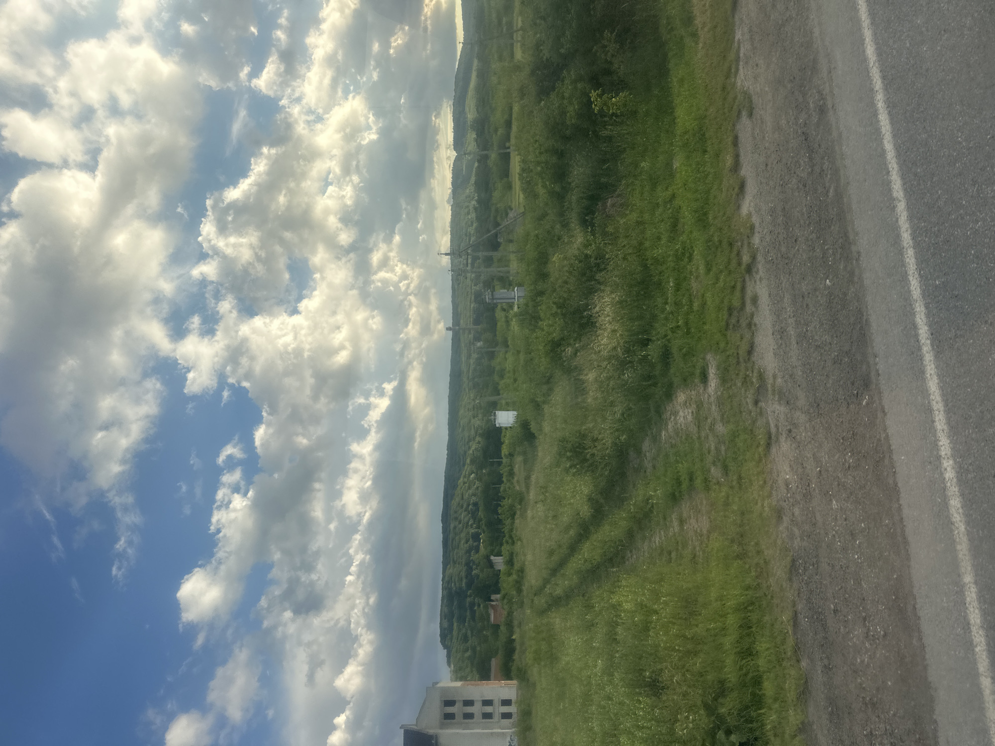 The image depicts a rural landscape with a partly cloudy sky. In the foreground, a paved road runs horizontally across the bottom of the frame, with a dirt track leading off into a grassy field. To the left, a multi-story building with a pale facade and dark windows stands partially in the frame. In the middle ground, the field is dotted with various objects including what appear to be electrical poles and boxes. Further in the background, rolling hills covered in dense green foliage stretch across the horizon. The lighting suggests it is either early morning or late afternoon. The location is identified as Zolochiv, Ukraine.