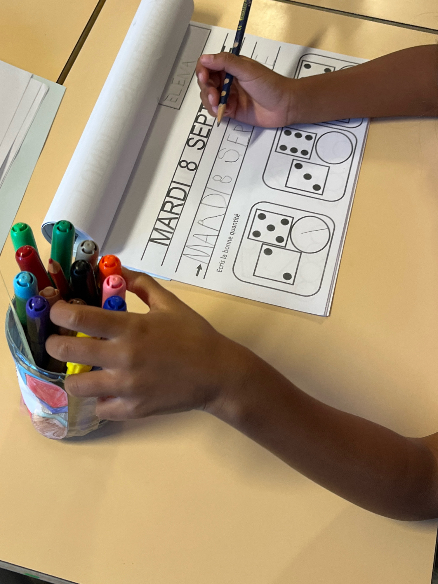 In La Fouillouse, France, a close-up, eye-level shot captures a child's hand holding a pencil, poised over a workbook. The workbook features a printed title in large, sans-serif letters that reads "MARDI 8 SEP" followed by "MAADI 8 SEP", with the second instance seemingly a handwritten repetition. Below this, a smaller instruction reads "Écris la bonne quantité". The page is divided into rectangular and circular compartments, some containing diagrams of dice with varying numbers of dots. One compartment shows a die with six dots, another with four, and a third with two. Another compartment contains a circle divided by a line. A cylindrical holder overflowing with colorful markers, including green, red, blue, orange, and purple, is positioned to the left of the workbook. The child's arm, with darker skin tone, extends from the right of the frame. The background consists of a light-colored surface, likely a desk, with a hint of a white paper roll on the left. The lighting is bright and even, suggesting an indoor setting.