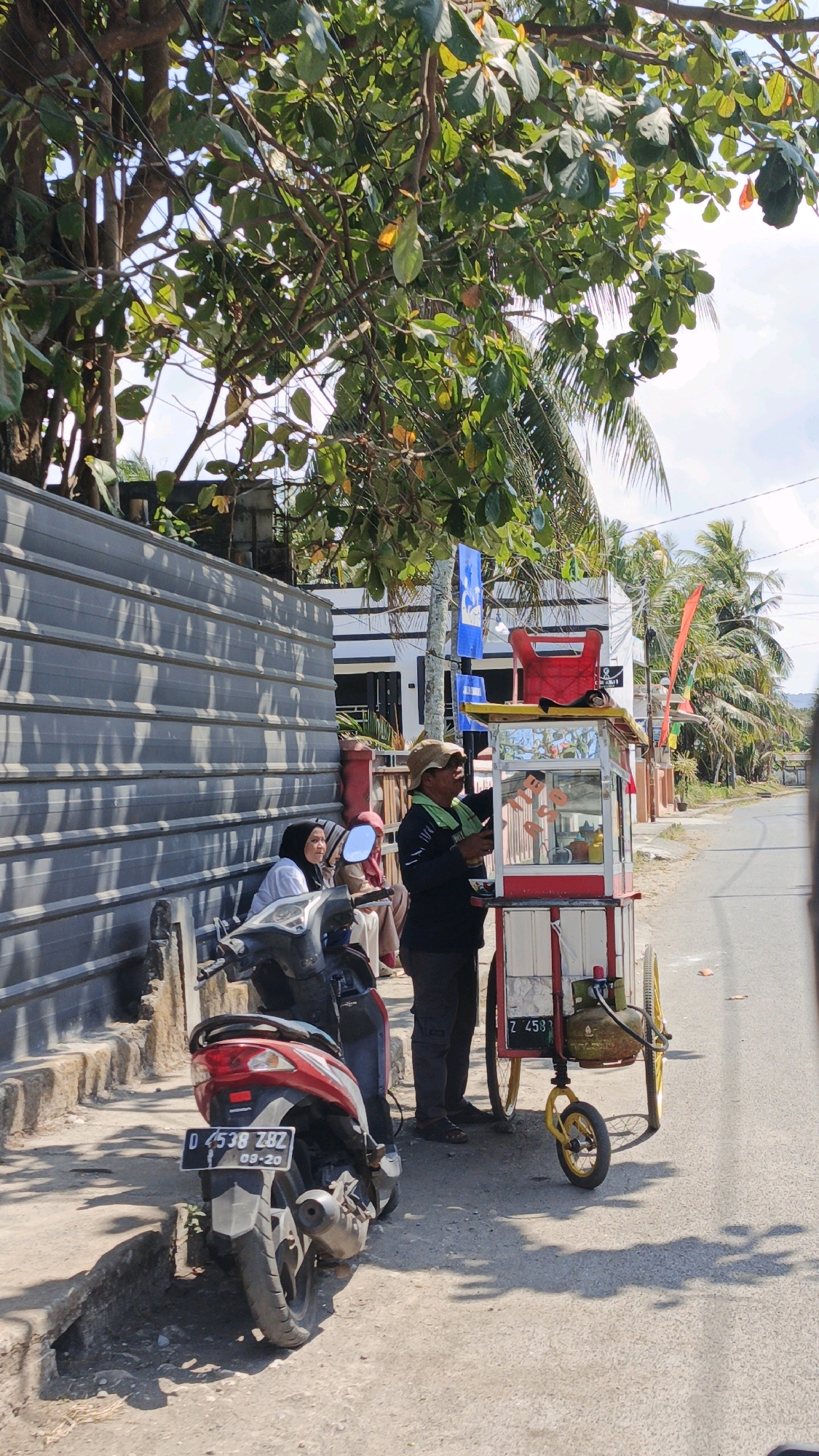 In Desa Pagandaran, Indonesia, a street scene unfolds under a canopy of leafy trees. A man wearing a light-colored hat and a green scarf over a dark shirt stands beside a mobile food cart. The cart, primarily red and white with yellow accents, displays a glass-enclosed section with food items, possibly donuts, and a gas cylinder attached to its base. The cart has two large yellow wheels and a smaller one at the front. To the left of the cart, a red scooter is parked. Two women, one wearing a white hijab and the other a light-colored headscarf, are seated on the edge of a gray corrugated metal fence. The fence runs along the side of a building with white siding and dark trim. The ground is paved with asphalt and dirt, with shadows cast by the trees and objects. The background shows more trees, including palm trees, and a street extending into the distance. The overall impression is a sunny day in a tropical setting.