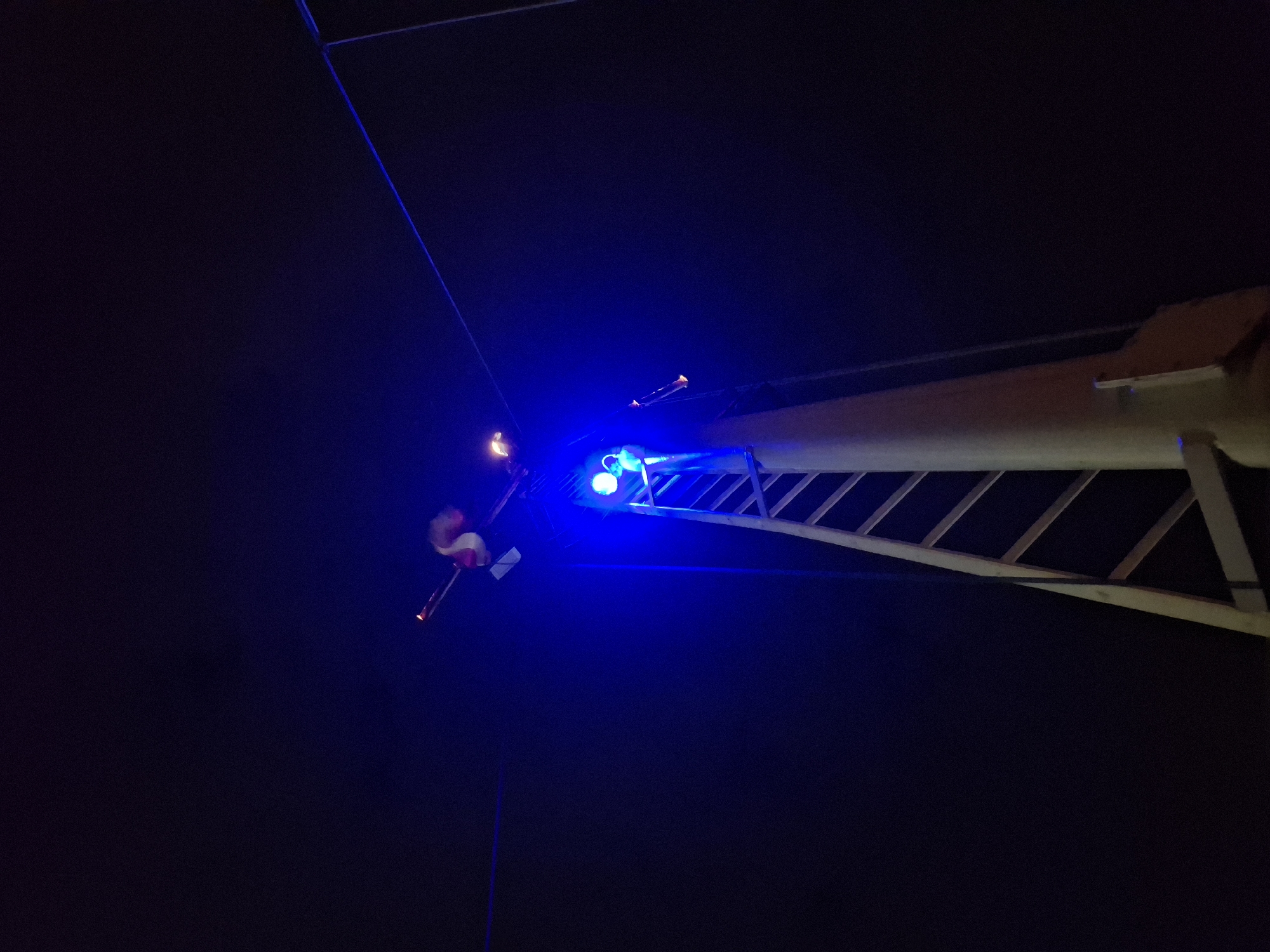 The image depicts a nighttime scene in an unknown city. A tall, beige-colored tower or structure is illuminated with bright blue lights. A series of ladder-like rungs extend along the structure's side. A figure in a red and white suit holding a lit torch is visible at the top. Thin cables extend into the dark sky above.