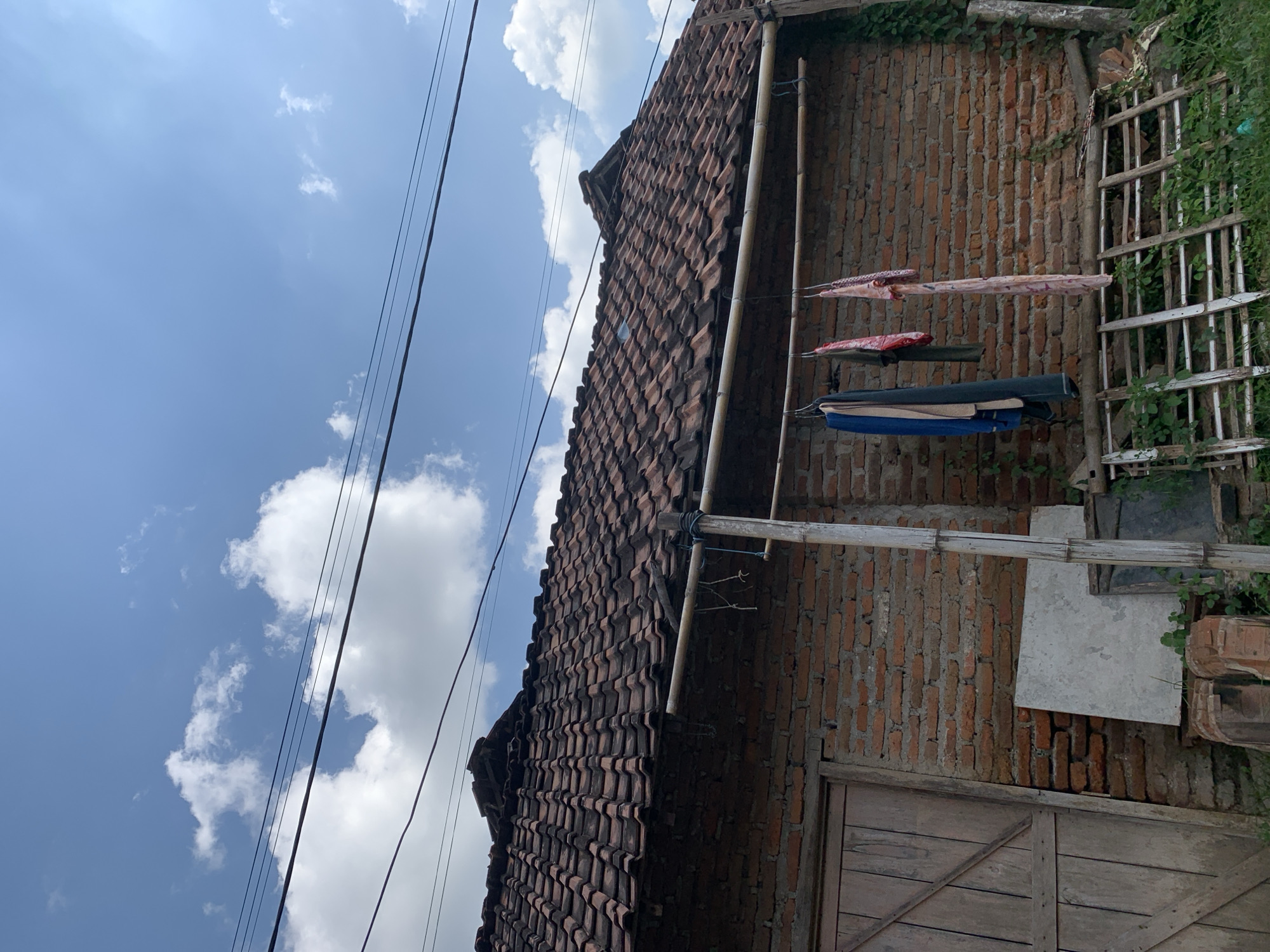 The image shows a brick house with a tiled roof under a blue sky with scattered clouds. Several articles of clothing are hanging on a clothesline in front of the house. A wooden door is visible on the left side of the house. In the foreground, there is a wooden rack and some greenery. The scene is outdoors in what appears to be a residential area. There are no people visible in the image. The overall impression is one of a simple, rural or suburban setting. The location is identified as Gulun, Indonesia.