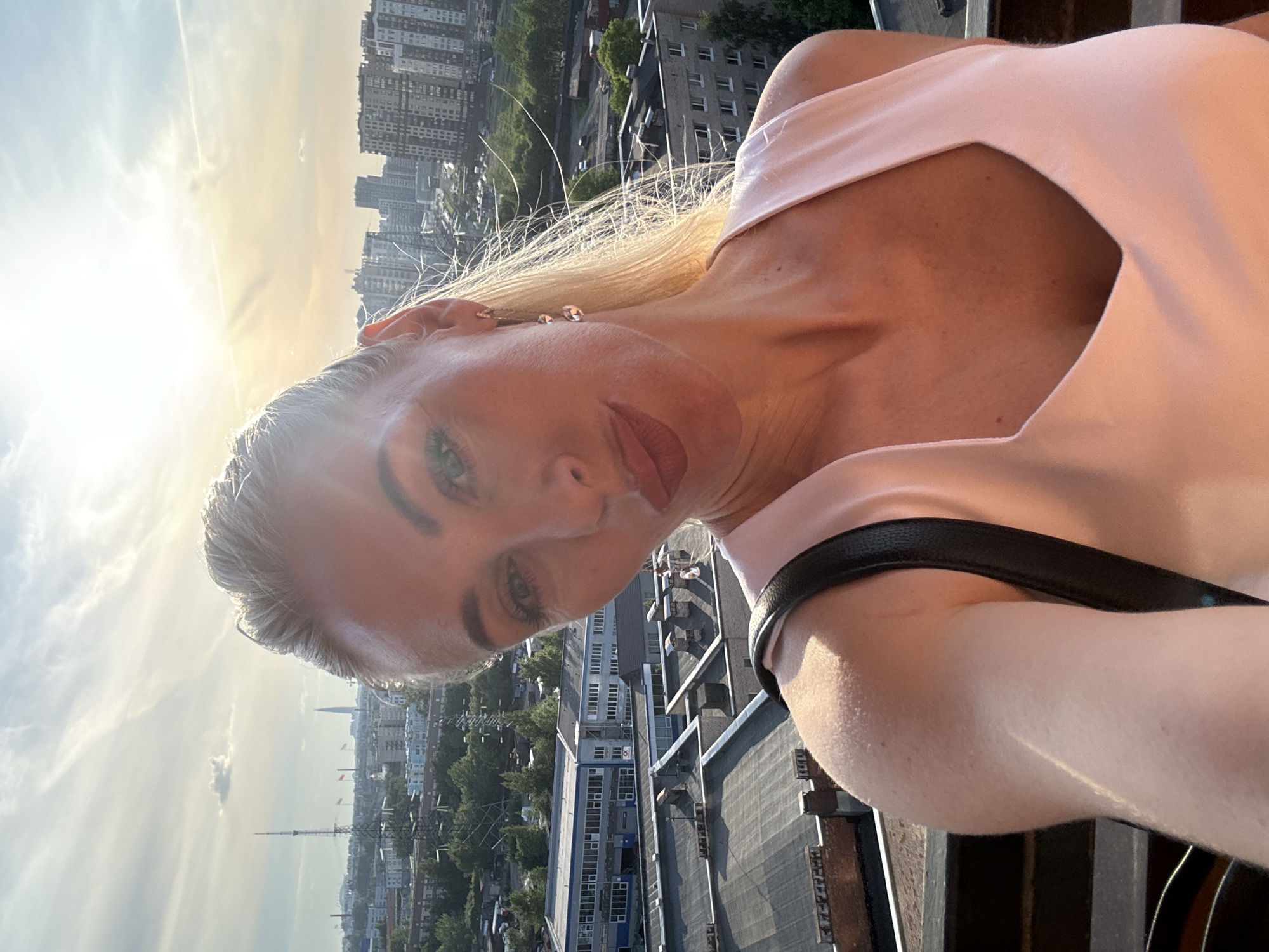 The image features a blonde woman taking a selfie from an elevated perspective overlooking the city of Saint Petersburg, Russia. She is positioned in the foreground, illuminated by sunlight from the upper right, and appears to be in her 30s-40s with a slight pout expression. She wears a light peach-colored sleeveless top with a V-neck and a black strap over her right shoulder, possibly from a bag. A silver-colored dangle earring is visible on her right ear. Her long blonde hair is styled back, with some strands catching the light. The background reveals a wide cityscape under a bright, partly cloudy sky. Distinct landmarks include the Saint Petersburg TV Tower on the left and the Lakhta Center skyscraper in the middle-distance on the right. The urban environment comprises various buildings, including industrial or commercial structures with dark flat roofs in the immediate foreground, and numerous residential apartment blocks of varying heights extending into the distance. Green areas, roads, and power lines are interspersed throughout the urban fabric.