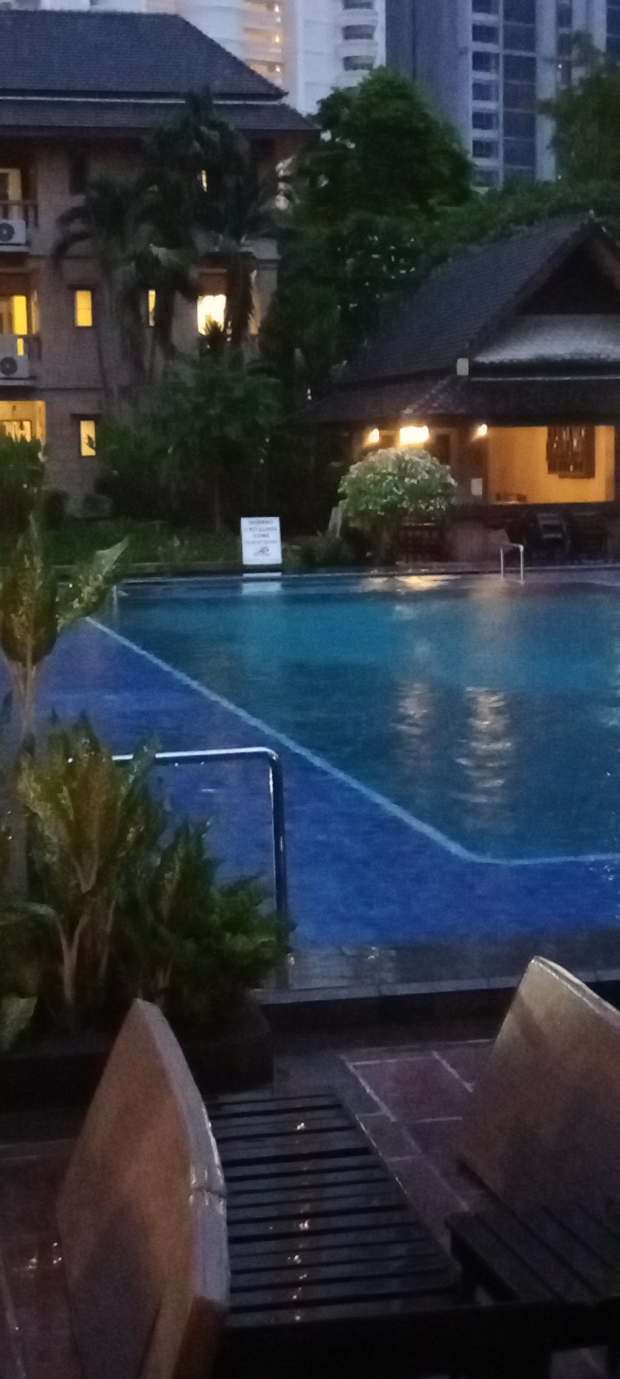 This image depicts an outdoor swimming pool area, likely at a resort or hotel, with a building in the background. The scene is set during twilight, with artificial lights from the building illuminating the area. The sky is a muted blue, suggesting the end of the day or a cloudy sky.
The pool itself is the central focus, with clear blue water reflecting the surrounding lights and trees. A blue tiled area surrounds part of the pool, with a metal railing visible along the edge. Lush green plants and trees are present, adding to the tropical ambiance. The building in the background appears to be a multi-story structure with several windows, some of which are lit. A smaller, traditional-looking building with a dark roof is also visible, possibly housing a facility or changing rooms for the pool.
In the foreground, partially visible, are lounge chairs made of dark wood and a pale, possibly stone, structure. The ground around the pool area appears to be wet, suggesting recent rain or ongoing maintenance. The overall mood of the image is peaceful and serene, inviting relaxation.
A small white sign with black text is visible near the edge of the pool. Although the text is too blurry to read in detail, it appears to be a warning or informational sign, possibly related to pool usage. The location context of Pattaya City, Thailand, reinforces the image of a tropical vacation spot. No people are visible in the frame.