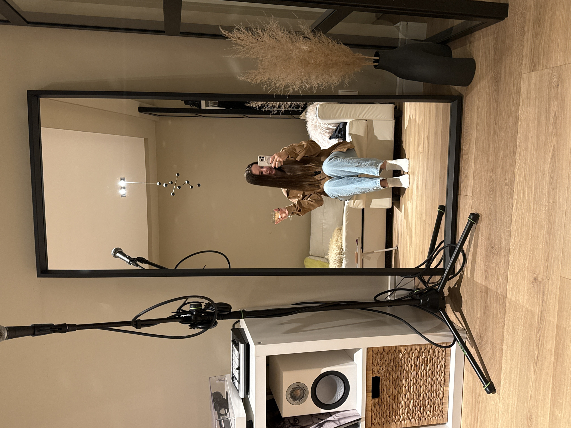 The image shows the reflection of a woman sitting on a white couch in a mirror. She is holding a phone in one hand and a glass in the other. The room appears to be a living space with beige walls and a light-colored wood floor. There's a black vase with dried flowers on top of the mirror frame. To the left, a microphone stand and a white shelving unit with a speaker and a woven basket are visible. The image provides no information to determine whether this location is in Barcelona, Spain.