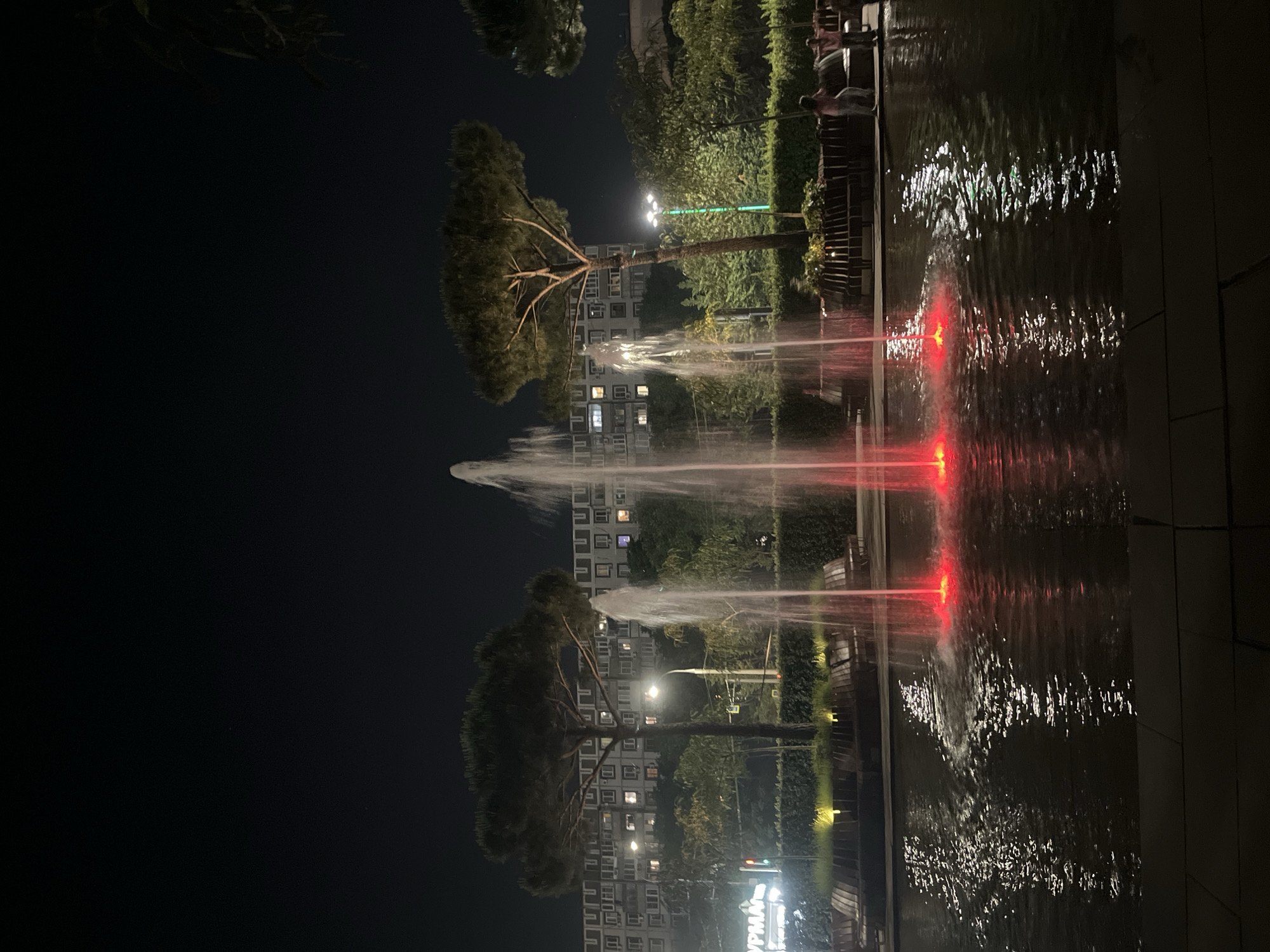 This is a night-time, vertical-orientation photograph taken in Krasnodar, Russia. The scene features a fountain with illuminated red jets of water, creating a vibrant contrast against the dark surroundings. The water's surface reflects the lights, adding to the dynamic visual effect.
In the background, a multi-story building with numerous lit windows suggests an urban residential area. Several trees, including a prominent pine tree with a distinctive canopy, are visible between the building and the fountain. A street lamp with white lights illuminates part of the scene, while another, possibly green, light is also present. Wooden benches and some greenery are arranged along the perimeter of the water feature, hinting at a public space designed for relaxation or recreation.
On the right side of the image, a few people are visible seated on a bench overlooking the fountain, seemingly enjoying the ambiance. The overall mood of the photograph is serene and captivating, with the interplay of light and water creating a sense of calm and beauty. The time of day is clearly night, indicated by the darkness and artificial lighting. There are no obvious weather cues, but the presence of water suggests it might be a humid or damp environment.
A partial view of a sign with Cyrillic text is visible at the bottom right, though it is too obscured to be fully deciphered. The image composition, with the fountain in the foreground and the building and trees in the background, creates a sense of depth and draws the viewer's eye through the illuminated elements of the scene.