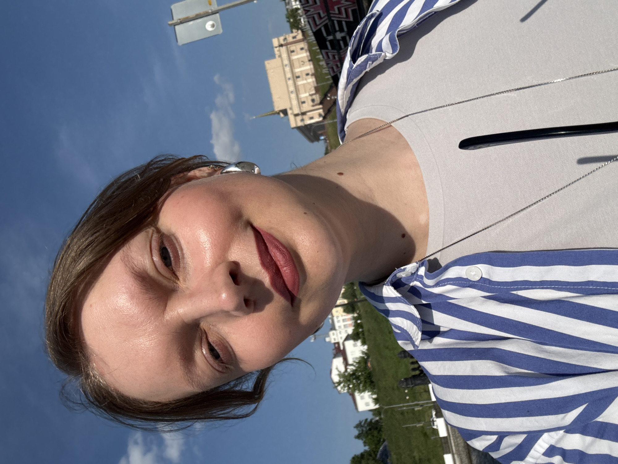 A person, presumably female, is shown in a selfie with a light-colored shirt and a blue and white striped shirt over it, wearing lipstick and a silver necklace. The background includes a blue sky and a building, most likely the Saint Michael's Cathedral in Izhevsk, Russia. Other buildings, trees, and a road sign are also visible.