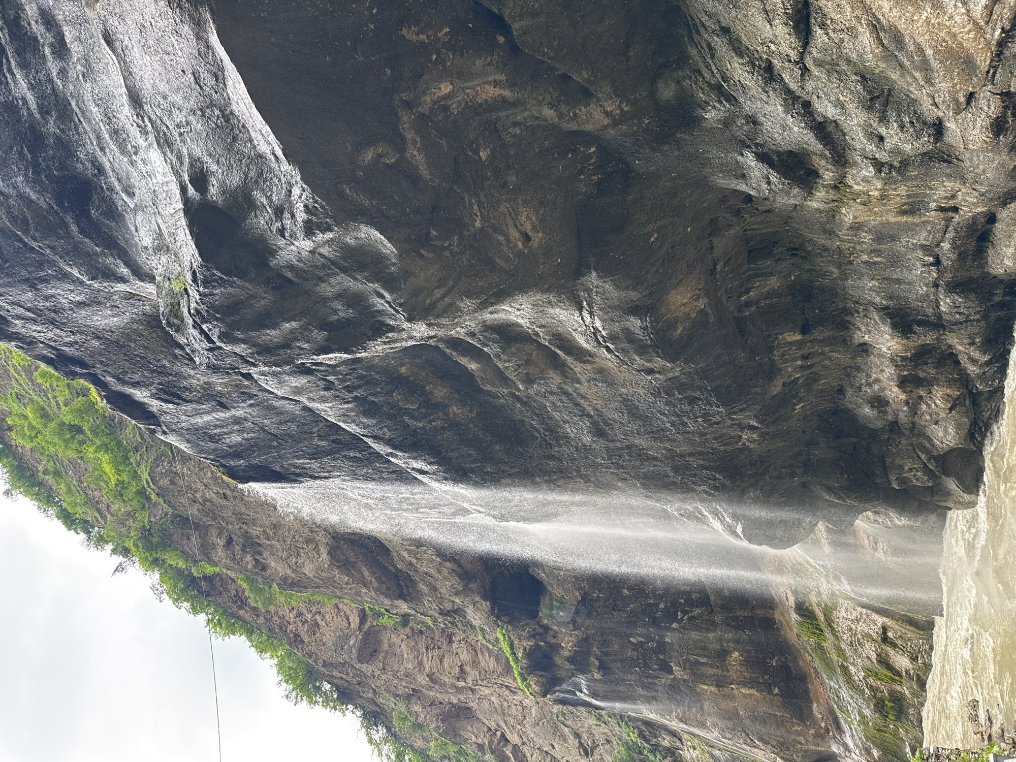 The image depicts a view of the Khushto-Syrt waterfall in Russia. A cascading waterfall, appearing as a thin stream of water, descends from a high cliff face on the left side of the frame. The cliff is a mix of rough, rocky textures and patches of green vegetation clinging to its upper ledges. The rock face is dark and wet, reflecting the light. To the right of the waterfall, a massive, dark, and uneven rock formation dominates the scene, with intricate patterns and textures on its surface. At the base of the waterfall, a turbulent, muddy river flows, with visible ripples and waves. The sky above is overcast and light. The overall impression is one of a natural, somewhat rugged, and powerful landscape.