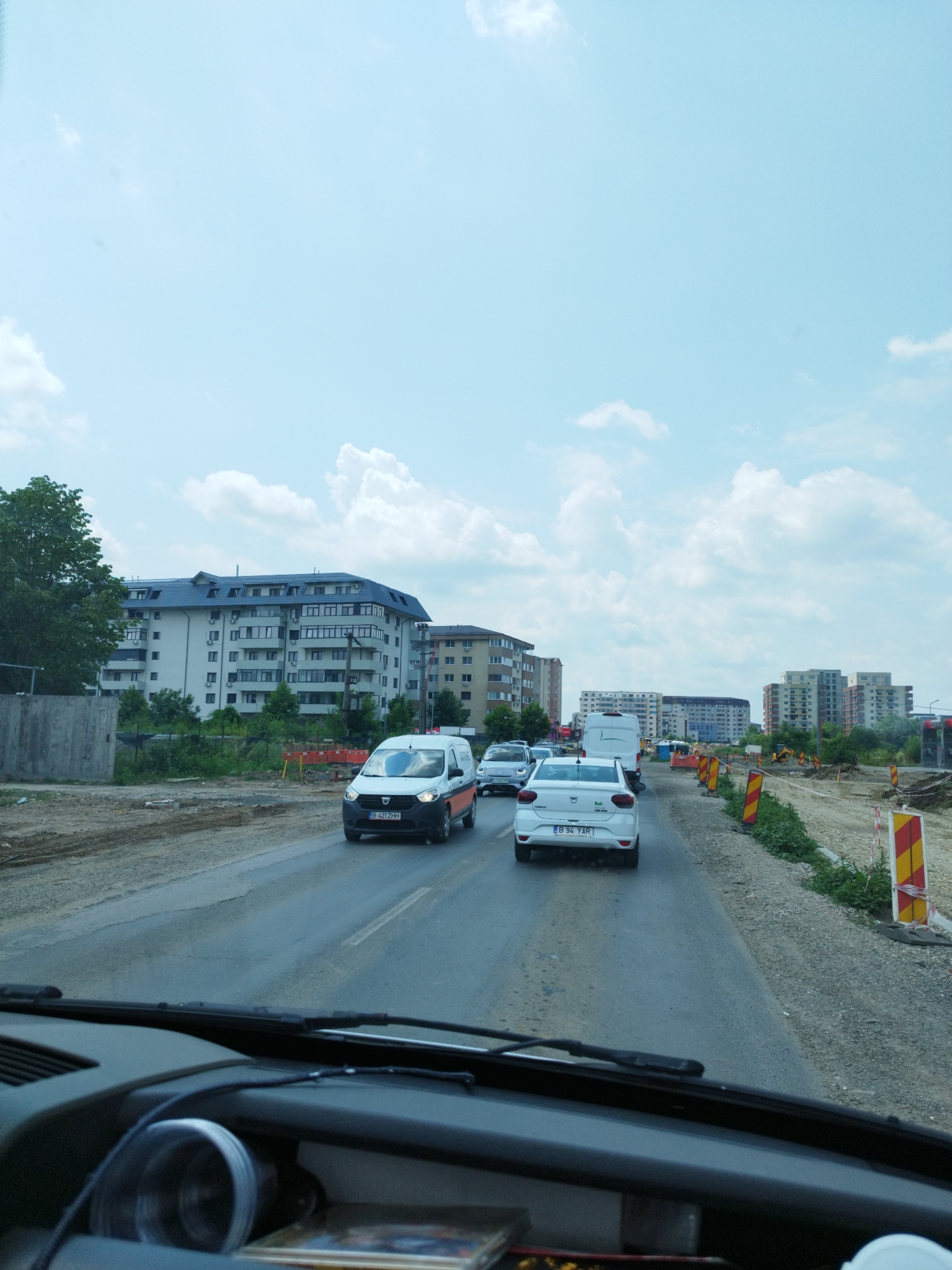 The image shows a view from inside a vehicle looking out at a street scene in Bragadiru, Romania. The street is a two-lane asphalt road with traffic. In the foreground, the dashboard and windshield wipers of the vehicle are visible.
On the road, several cars are present. A white van with the license plate B 421ZHH is in the middle lane. Ahead of the van is a white Dacia car with the license plate B 94 YAR. Further ahead, there are other vehicles, including another white van and a white car.
The environment indicates construction is taking place. The right side of the road is lined with construction barriers, including yellow and red striped traffic cones and other orange construction equipment. The left side of the road features a tall, multi-story residential building with a dark roof and numerous windows. Adjacent to this building is a concrete wall. Beyond the buildings, the sky is partly cloudy and blue. The overall impression is a suburban or urban area undergoing road work.