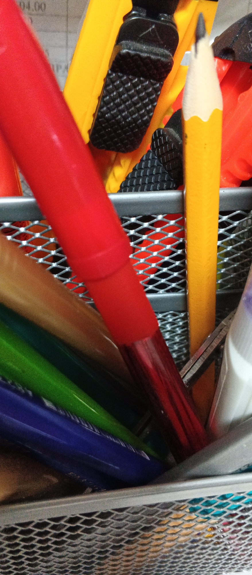 The image displays a close-up view of a metal mesh desk organizer filled with various writing instruments. Dominating the foreground is a thick, red marker, its surface appearing smooth and shiny. To its right, a yellow pencil with a sharpened graphite tip is prominently visible. Behind these, other items are partially obscured, including what appears to be a yellow and orange stationery item with a black textured grip, possibly a craft knife or box cutter. Several colored pencils or markers are visible at the bottom, including green and blue. The organizer itself is made of silver-colored metal mesh with a diamond pattern. The background consists of a surface with faint text, possibly a document or desk mat. The lighting suggests an indoor environment. There are no people or animals present. The image provides no information about the city of Smila, Ukraine, or any specific location within it.
