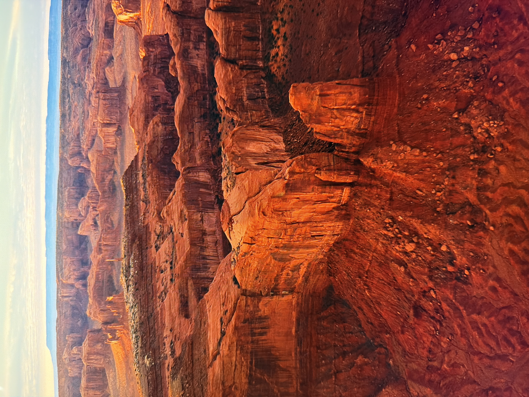 The media file depicts a vast, arid canyon landscape bathed in warm, golden light, suggesting either sunrise or sunset. Towering rock formations, characterized by their layered sedimentary structure and vibrant red and orange hues, dominate the scene. The scale of the canyon is emphasized by the distant, hazy horizon line, where a pale blue sky meets the rugged terrain. Small patches of vegetation, appearing as dark green or brown clusters, cling to the cliff faces, adding subtle texture and color variation. The rocks themselves exhibit intricate patterns of erosion, with sharp edges, deep crevices, and smooth, curved surfaces. Scattered across some of the rock faces are small pebbles and debris, indicating the natural processes of weathering. The overall impression is one of immense natural beauty, geological grandeur, and a sense of serene isolation. There are no visible people, objects, or text in the image. The location context of Kyiv, Ukraine, is not reflected in the visual content of the image.