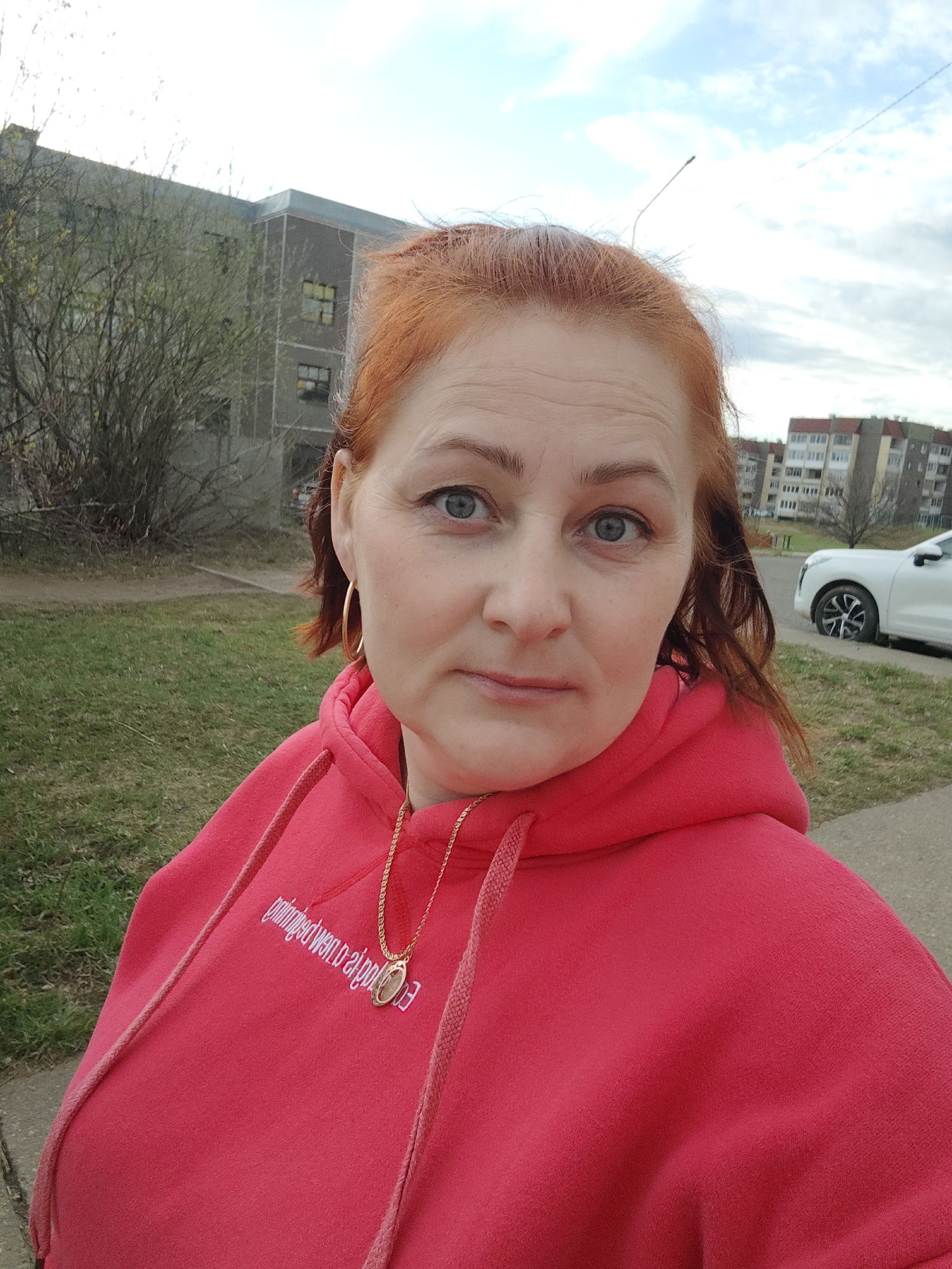 A medium shot captures a woman outdoors in what appears to be a residential area of Markovskiy, Russia. She is centrally framed, looking directly at the camera with a neutral expression. Her hair is a reddish-brown, and she has light blue eyes. She is wearing a bright red hooded sweatshirt (hoodie) with the hood down. A gold chain necklace with a circular pendant rests on her chest, and she has gold hoop earrings.
In the background, to her left, are bare-branched trees or bushes, partially obscuring a multi-story, grey apartment building. To her right, another apartment building with a lighter facade and reddish roof tiles is visible further in the distance. A white car is parked on the side of a paved area in the right background. The ground is covered with green grass and a paved path runs along the bottom of the frame. The sky is partly cloudy, suggesting it is daytime with diffused natural light.
On the front of her red hoodie, there is white text that appears reversed and partially obscured by her necklace and the drawstring. The text is difficult to decipher but seems to include phrases like "supporting Drew" followed by other letters.