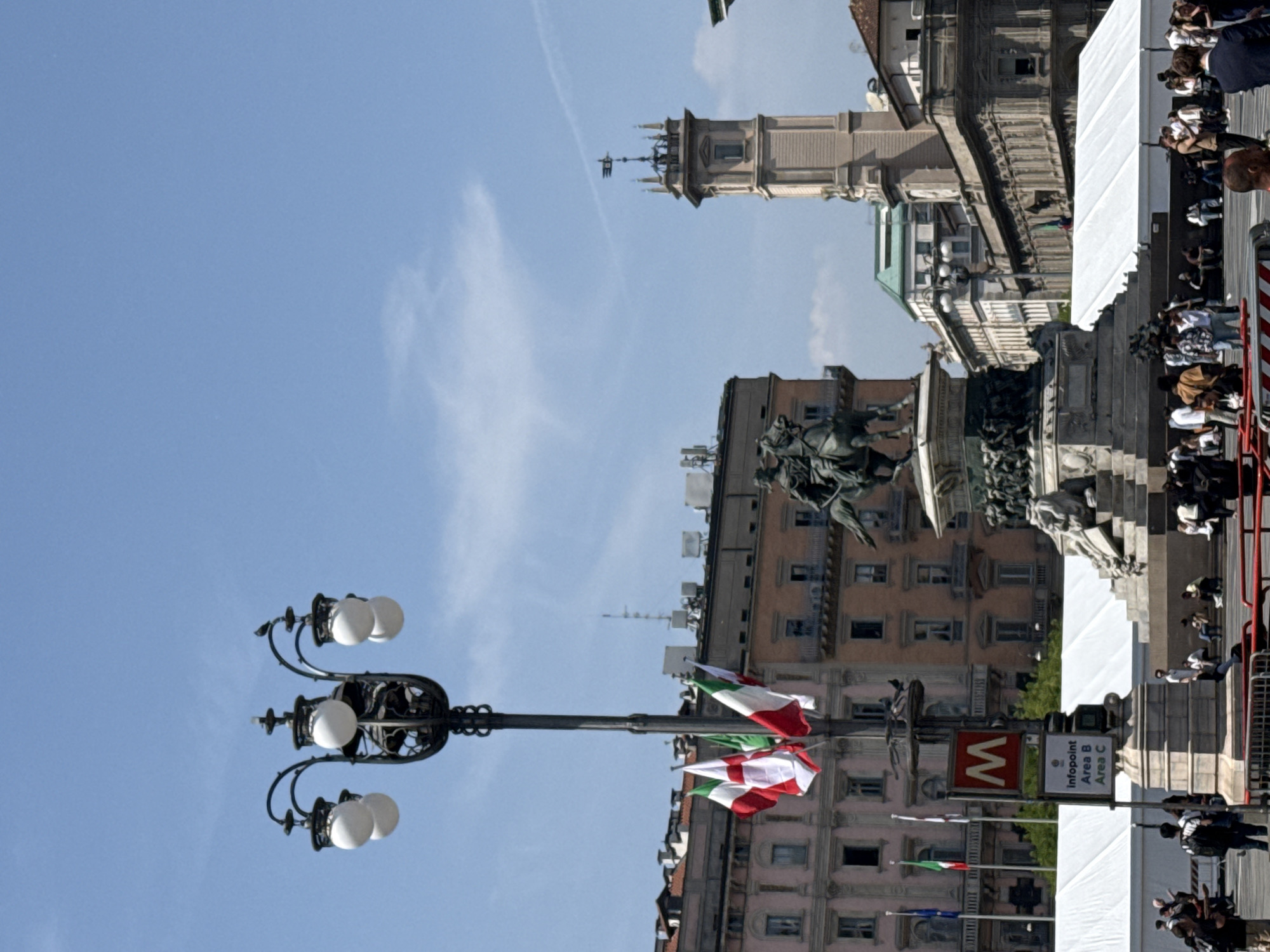 This image, taken in Milan, Italy, depicts a bustling public square or street scene under a clear blue sky with a few wispy clouds, indicating daytime. In the foreground, an ornate black metal lamppost, featuring multiple white globe lights, stands prominently. Several Italian flags (green, white, red vertical stripes) are attached to this lamppost, along with another flag displaying horizontal red, white, and green stripes.
To the right of the lamppost, a red "M" sign, characteristic of the Milanese Metropolitana (metro system), is visible. Below it, a white rectangular sign reads "Infopoint Area B Area C," referencing Milan's traffic restriction zones.
The background features several historic buildings with intricate architectural details. A tall, slender tower with a spire and a weather vane rises above the other structures. In the midground, a large equestrian statue, possibly of a historical figure, is situated on a stone pedestal, surrounded by other statues and reliefs.
Numerous people are gathered on the right side of the image, along a wide paved area and near temporary white tent-like structures. Red and white striped barriers suggest crowd control or demarcation for an event. The individuals appear to be observing or participating in a public gathering or celebration, given the flags, infopoint, and general setup. The activity suggests a vibrant, organized event in a prominent city location.