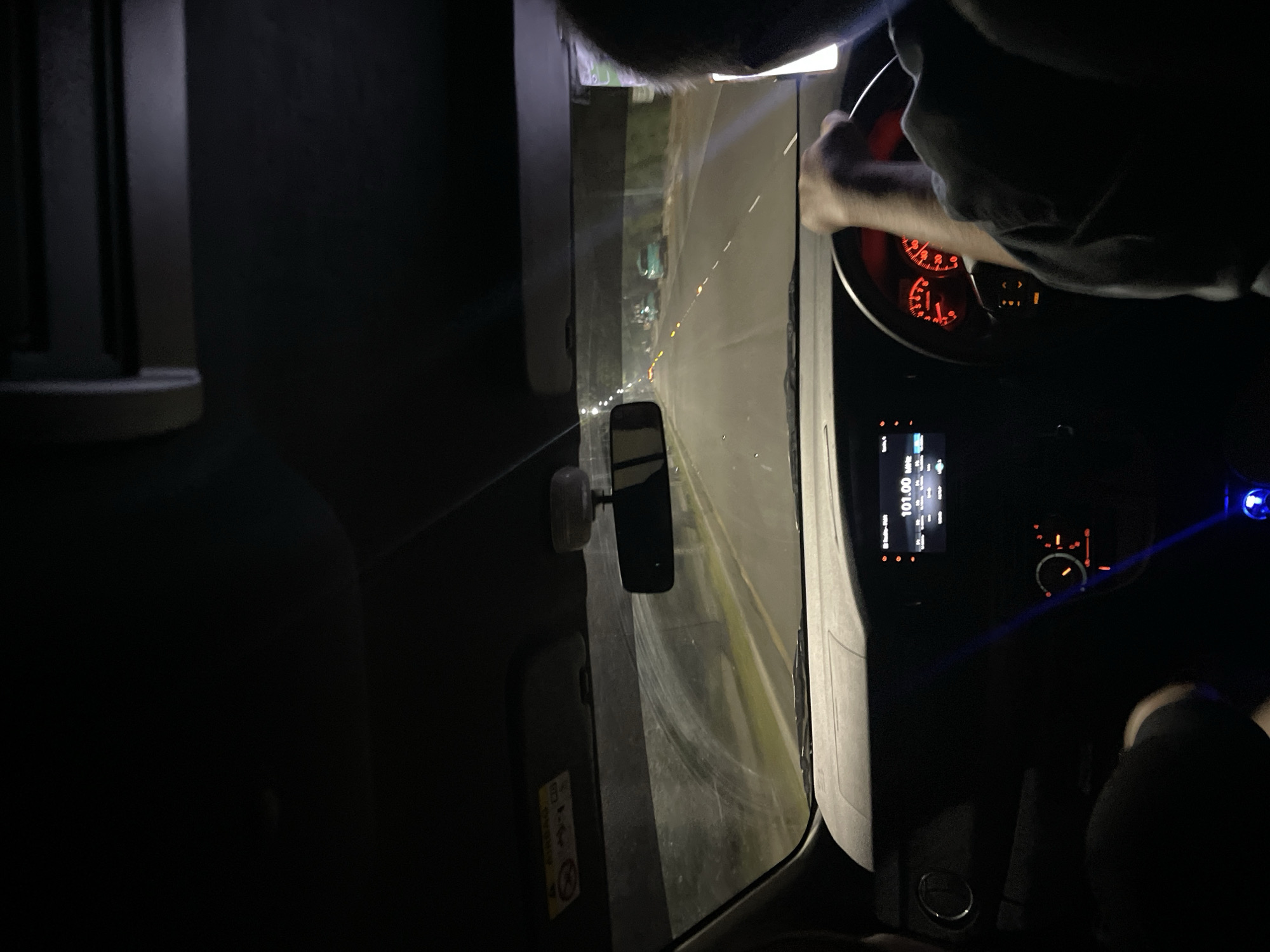 The image depicts the interior of a vehicle at night, facing forward. The driver's hands are visible on the steering wheel. Through the windshield, a road stretches into the distance with other vehicles visible. The dashboard is illuminated, displaying a digital display reading "101.00." The speedometer and other gauges are lit. To the left, a side mirror is visible. The location is possibly in North Jakarta, Indonesia.