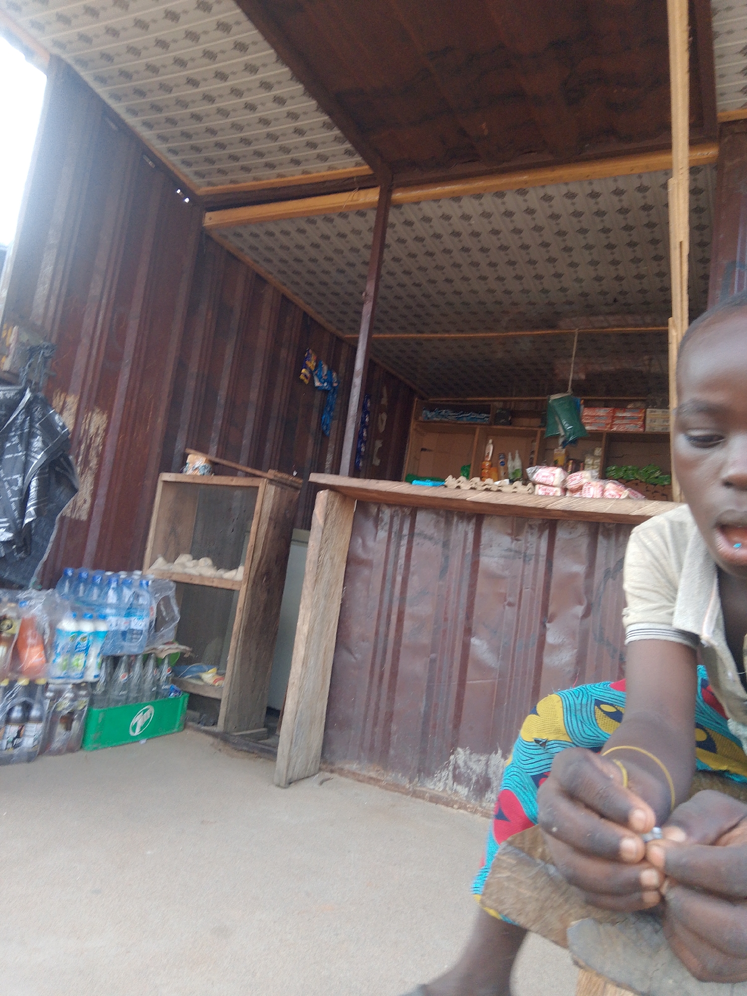 This image depicts a roadside stall in Jalingo, Nigeria. A young person, possibly a child, is seated in the foreground, their hands actively engaged with something small and metallic, perhaps a piece of jewelry or an accessory. They are wearing a vibrantly patterned blue and yellow fabric. The stall itself is constructed from corrugated metal and wood, with shelves stocked with various goods, including bottled water and what appear to be food items or packaged products. A black plastic bag hangs on the left side of the stall, and a wooden shelf unit is visible within the stall's structure. The ground is a light-colored, sandy surface, suggesting an outdoor, possibly dusty environment. The lighting indicates it is daytime.