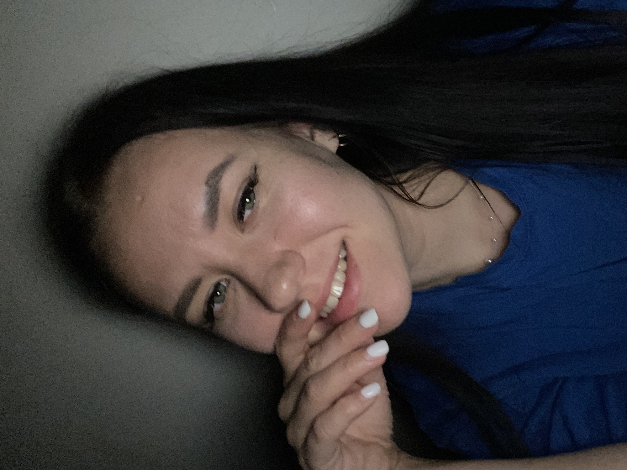 The image, presented sideways, features a woman with long, dark hair, fair skin, and striking green eyes, smiling broadly towards the camera. Her head is tilted, giving the impression she is lying down or reclining. She is wearing a short-sleeved blue top. Her left hand, adorned with white nail polish, is gently placed over her lower face, near her mouth, contributing to her warm, possibly slightly shy or playful, expression.
Notable details include a small hoop earring visible on her right ear and a delicate silver chain necklace around her neck. The lighting is somewhat dim, suggesting an indoor setting with low ambient light, which adds a soft, intimate feel to the portrait. The background is indistinct: a plain dark grey surface is visible behind her head, and a blue fabric, possibly part of a bed or sofa, is behind her body. There is no visible text. The overall mood is cheerful and candid. No specific location cues for Chaikovsky, Russia are visible within the image itself.