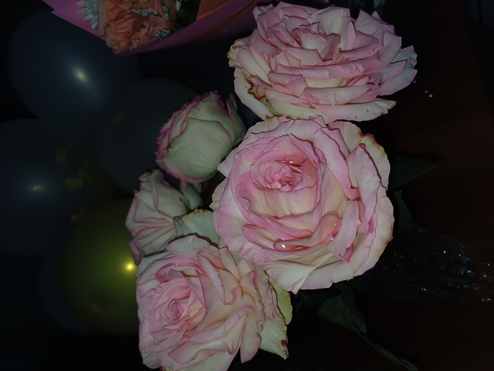 The image shows a close-up of a bouquet of light pink roses, some with dew drops on their petals, against a dark background. In the upper portion of the image, out of focus, are several balloons of various colors, including green, dark blue, and grey, one of which has yellow speckles. A hint of a pink and orange floral wrapping is visible on the right side. The scene appears to be indoors, with the dark background suggesting a dimly lit environment. There are no people visible in the image.