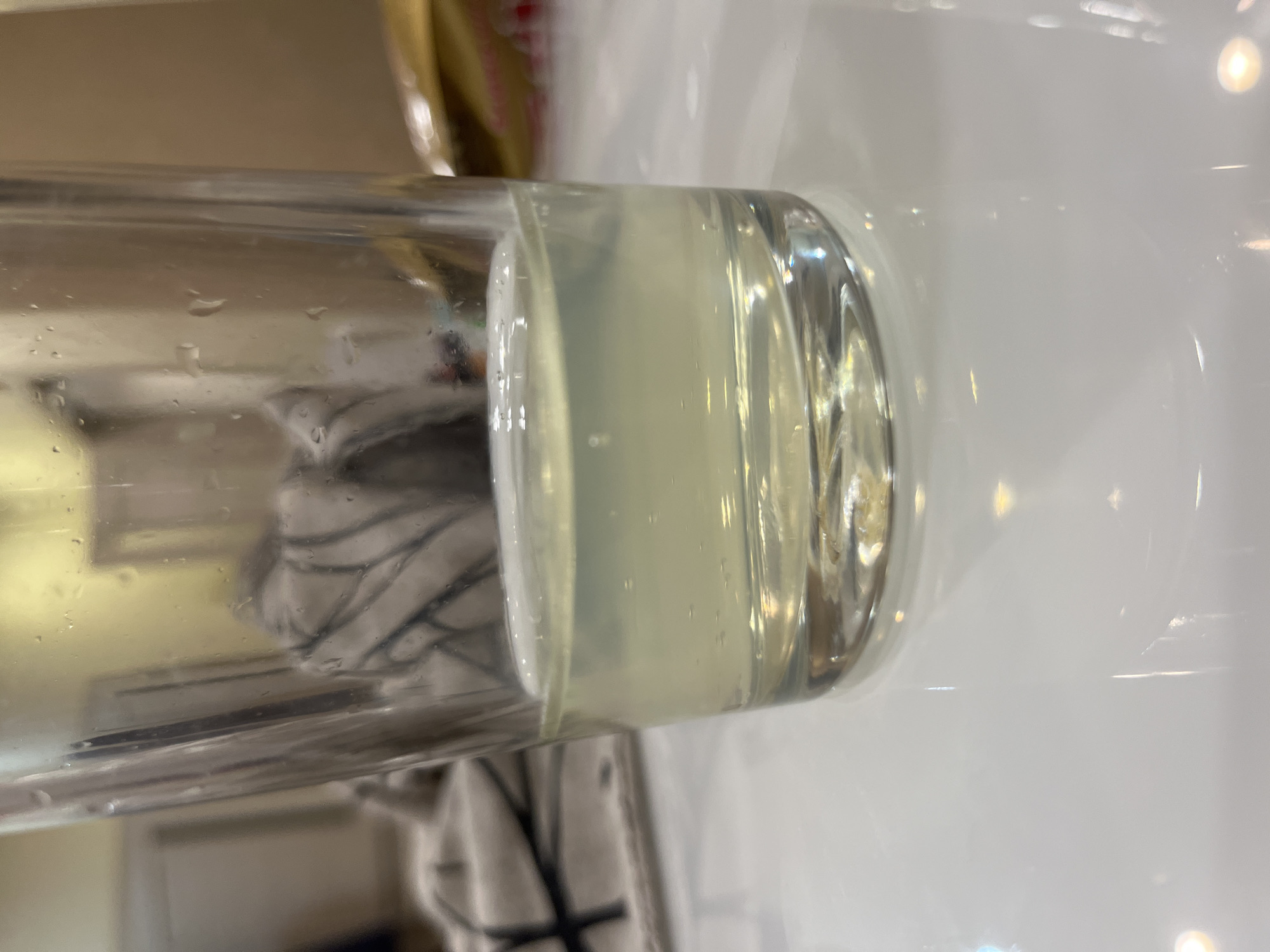 The scene is an interior view within the city of Tyumen, Russia, depicting a clear glass tumbler containing a light yellowish-green liquid, approximately one-quarter full. Small air bubbles adhere to the interior walls of the glass and are suspended within the liquid. The exterior surface of the glass exhibits numerous water droplets, indicative of condensation or recent contact with water. The glass rests on a highly reflective white surface, likely a countertop or table, which shows specular reflections of overhead lighting. Through the transparent glass, the background reveals a blurred light-colored fabric with a dark, possibly abstract, pattern on the left, and a muted, darker beige or brown wall segment further to the right. An indistinct yellow light source or reflection is visible in the upper background. No individuals are present in the frame.