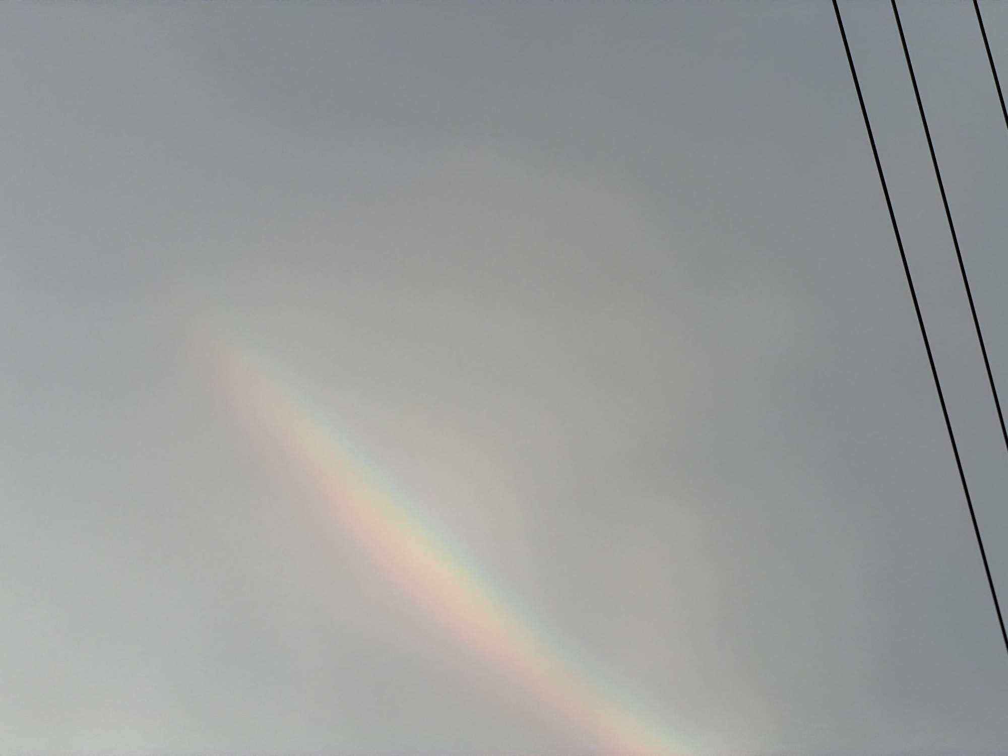 The image displays a sky primarily composed of light gray, suggesting overcast or hazy weather conditions. A faint rainbow is visible, arcing diagonally across the lower left section of the frame. Its colors, though subtle, include hues of red, orange, yellow, and green. In the upper right corner, three dark, thin lines, likely utility wires, stretch diagonally downwards across the sky. These lines are parallel and evenly spaced.
No people or specific activities are depicted in the scene, which captures a natural phenomenon alongside human infrastructure. The presence of the rainbow indicates that sunlight is interacting with water droplets in the atmosphere, suggesting recent rain or high humidity despite the cloudy sky. The wires point to an urban or semi-urban setting within Phan Rang Ward, Vietnam. There is no visible text within the image.