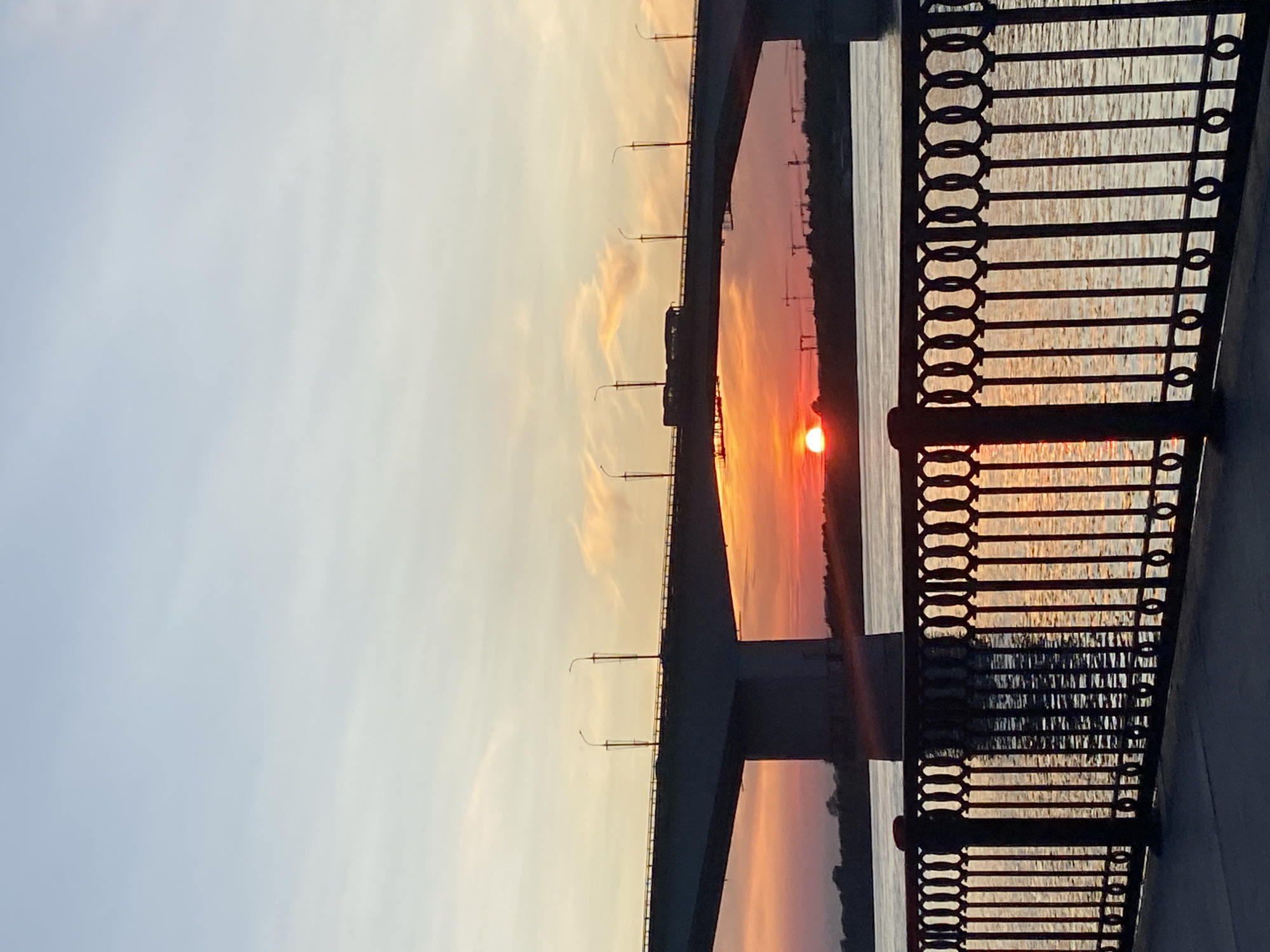 The image depicts a sunset over the Kama River in Perm, Russia, viewed from the embankment. A large concrete bridge, likely the Kama Bridge or the Imperial Bridge given its size and location, spans the river. The sun, a vibrant orange disc, is setting behind the distant shoreline, casting a warm glow across the sky and water. Silhouettes of industrial cranes are visible on the far bank, suggesting an industrial area. A bus is seen crossing the bridge, its form a dark silhouette against the colorful sky. The foreground features a black ornate metal fence that runs along the embankment, with its reflection visible in the rippling water. The sky transitions from a pale blue at the top to hues of orange, pink, and red near the horizon. The water is calm, reflecting the colors of the sunset and the bridge structure.