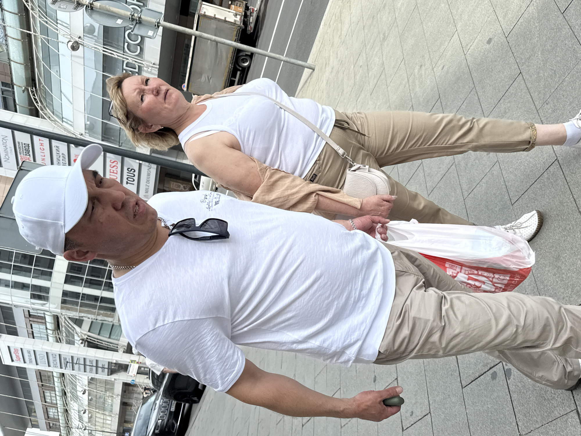 In Moscow, Russia, a man and a woman are walking down a sidewalk. The man, wearing a white t-shirt, white baseball cap, and beige pants, is holding a green object in his left hand and a white plastic bag with red lettering in his right. The woman, wearing a white tank top, a beige cardigan draped over her shoulders, and beige pants, has a white crossbody bag. They are walking next to each other, with the man slightly ahead of the woman. In the background, a modern building with a glass facade is visible, along with street signs and a truck. The sidewalk is paved with gray rectangular tiles.