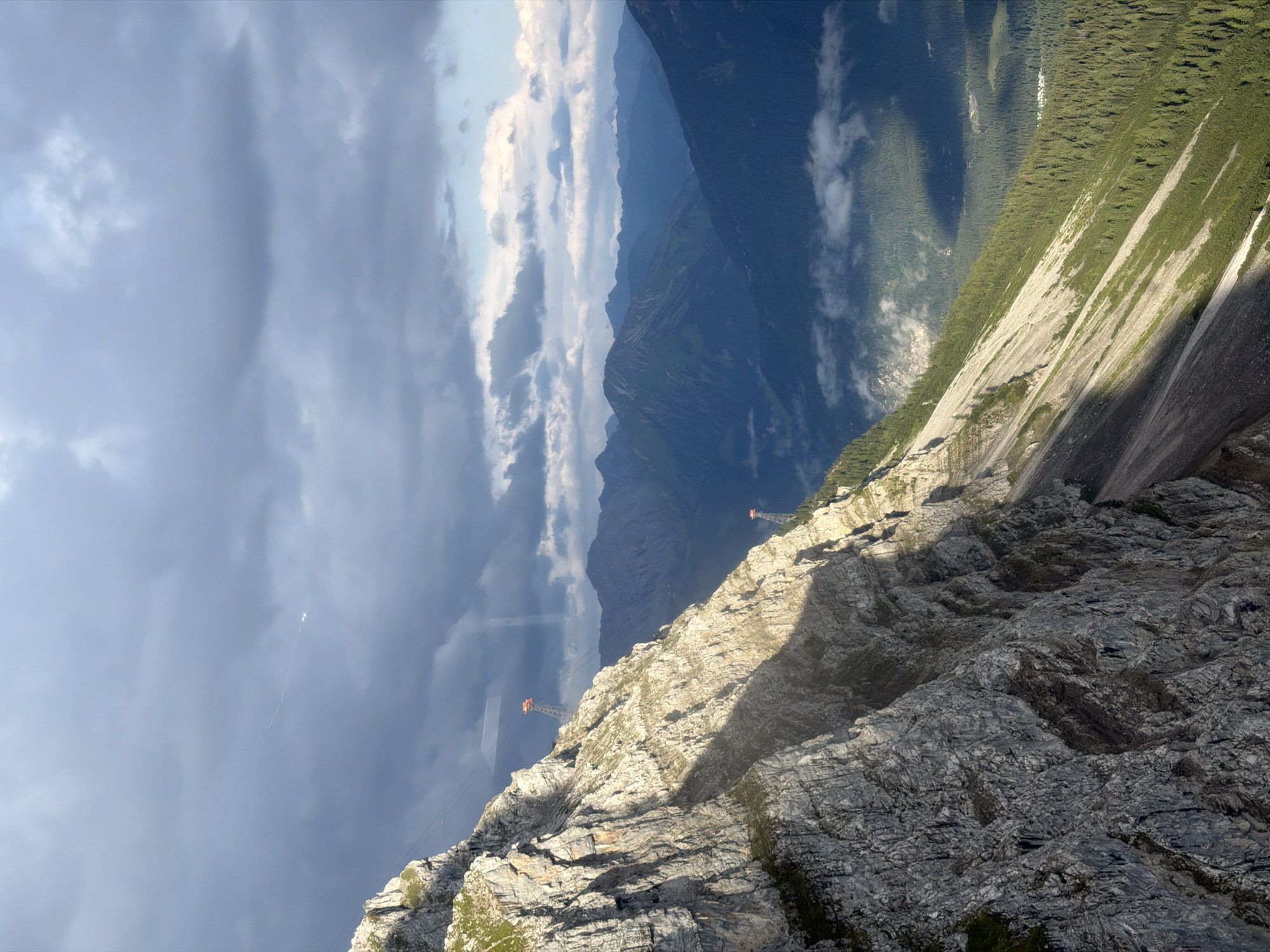 The image shows a dramatic mountain landscape, likely taken from a cable car, with a steep, rocky cliff face dominating the left side. Sunlight casts a strong shadow across the cliff, highlighting its rugged texture and the sparse vegetation clinging to its ledges. In the background, a series of lush, forested mountain ranges recede into the distance, with some low-lying clouds drifting through the valleys.
Two red cable car towers are visible on the mountainside, indicating a transportation system is in place. One tower is higher and closer to the viewer, while the other is further down the slope. The sky above is a mix of dark, dramatic storm clouds and brighter, scattered clouds, suggesting variable weather conditions. The presence of cable car infrastructure suggests this is a popular tourist destination for hiking and mountain excursions, possibly in the Alps. While the image does not definitively place the location in Grainau, Germany, the description of mountainous terrain and cable car systems is consistent with areas like the Zugspitze, Germany's highest peak, which is accessible via cable car and located near Grainau.