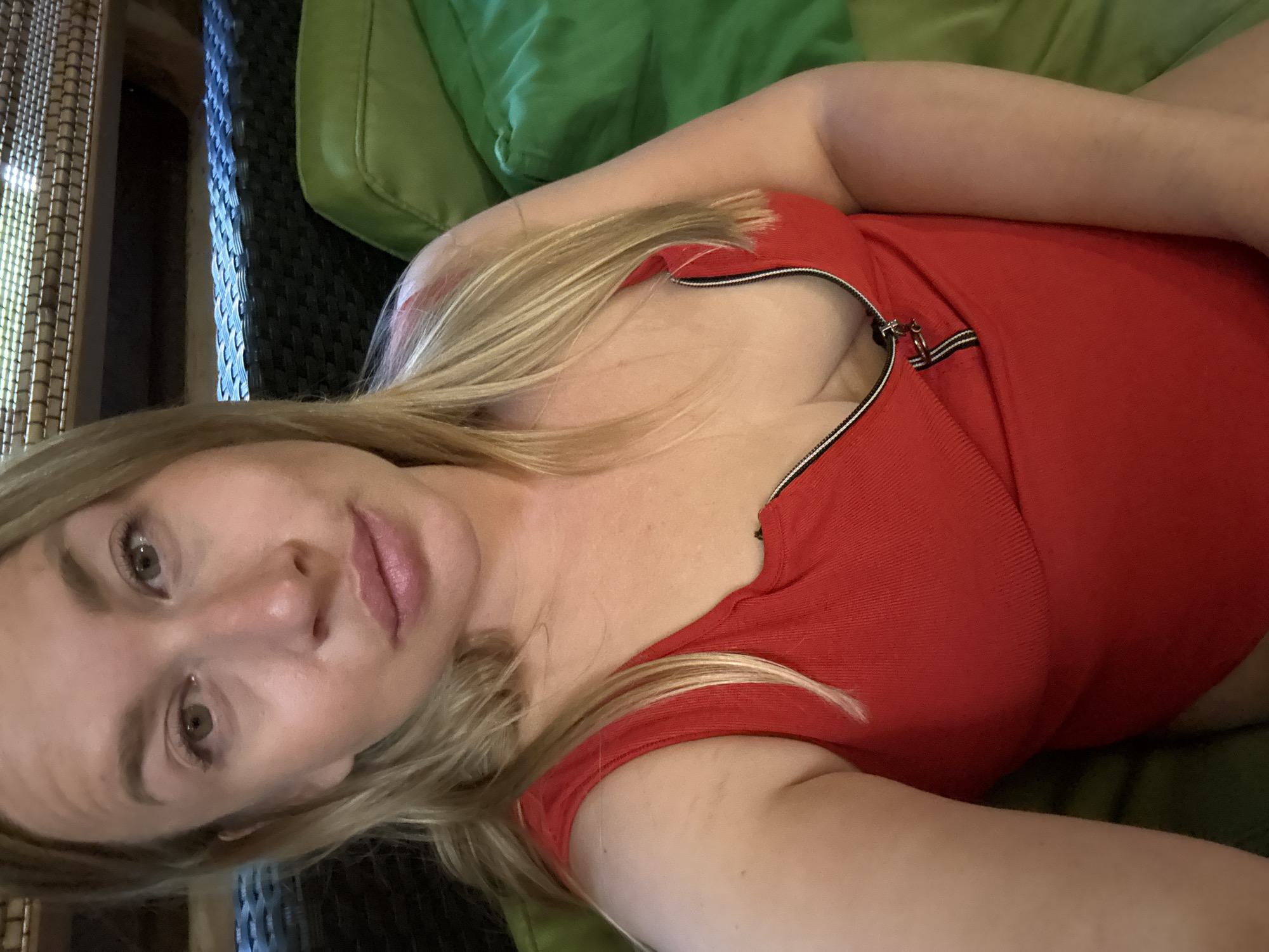 A blonde woman with light skin and a light complexion is taking a selfie indoors. She is wearing a red one-piece swimsuit with a zippered neckline, partially unzipped to reveal her cleavage. Her hair is parted in the middle and falls past her shoulders. She is sitting on a green couch. The background shows woven textures of what appears to be outdoor furniture, possibly on a patio or deck. The lighting is dim, with a light source from above. No specific city or location details are present in the image.