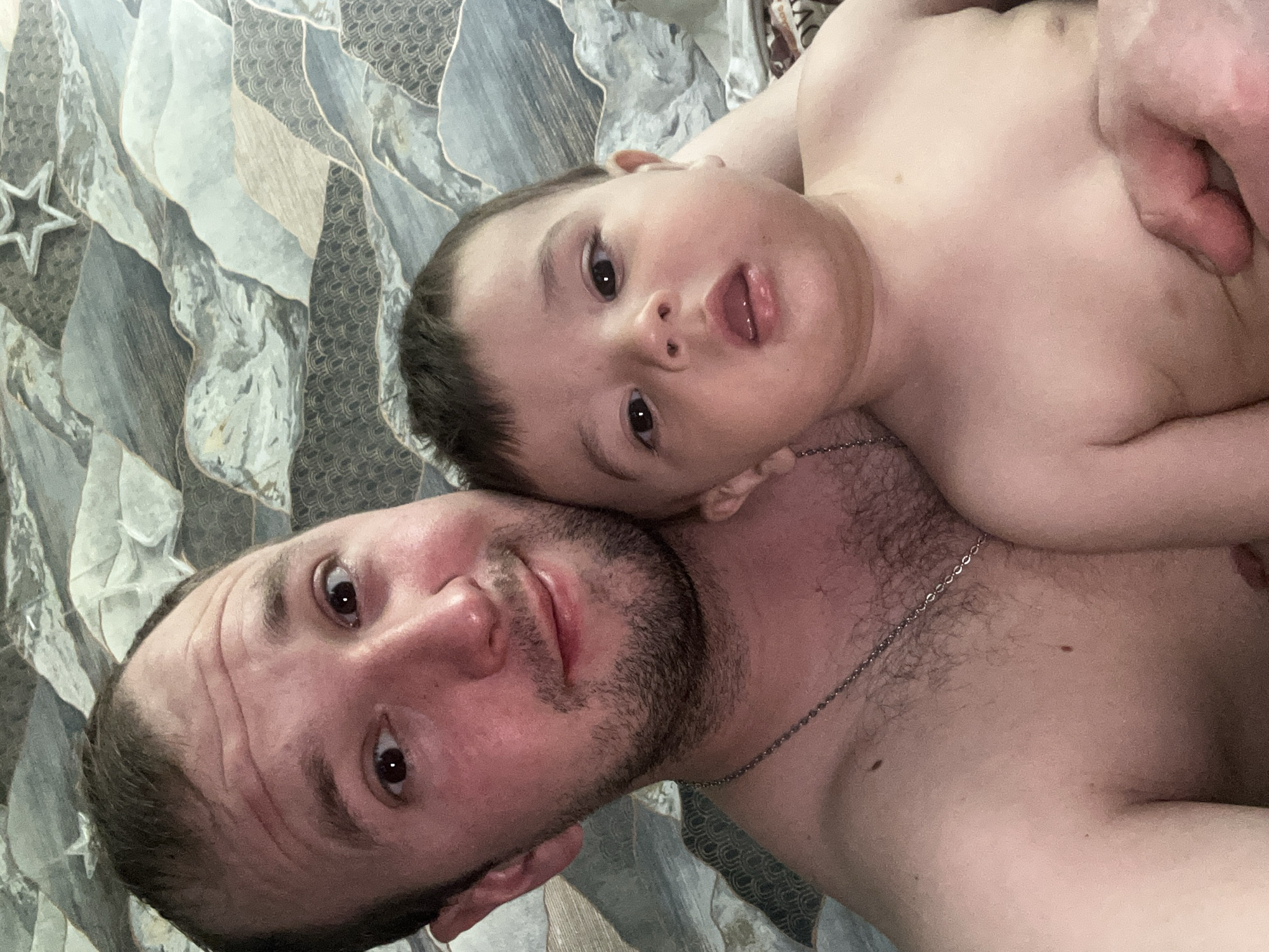 A close-up photograph shows a shirtless man and a young child lying on what appears to be a patterned bedspread. The man has short brown hair, a beard, and is looking directly at the camera. The child is lying on the man's chest, also looking at the camera. A silver chain is visible around the man's neck. The background consists of a gray and white patterned material. The image was taken in Chaikovsky, Russia.