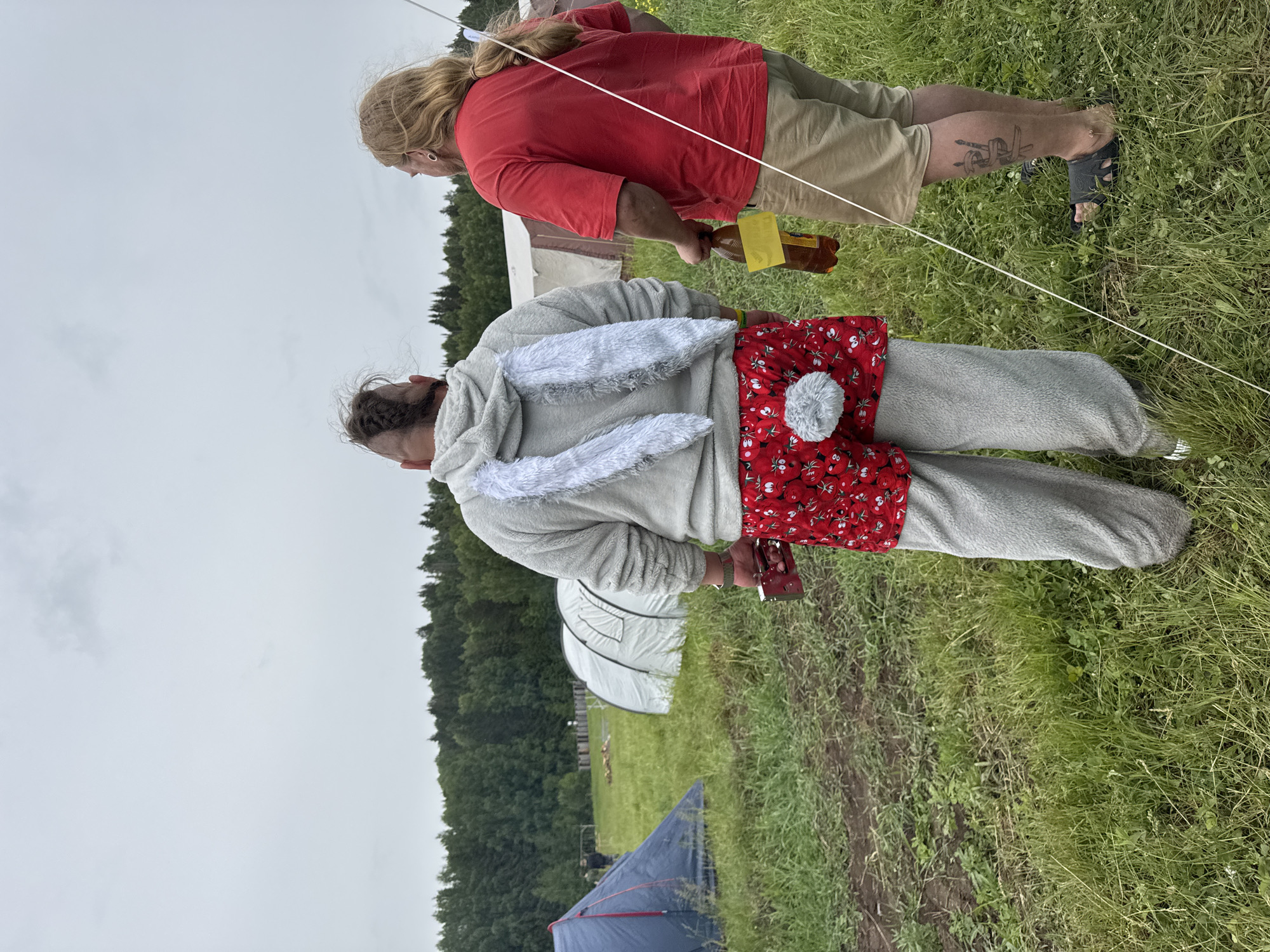 This image captures an outdoor scene at what appears to be a casual gathering or campsite in Большая Кивара, Russia. The perspective is slightly elevated, looking down on two individuals standing in a grassy field under an overcast sky.
In the foreground, a person is seen from the back, wearing a distinctive grey bunny-themed kigurumi or pajamas. This outfit features large, white fluffy ears on the hood and a round, fluffy grey tail on the back. Over the kigurumi, a red apron or skirt with a white floral or fruit pattern (possibly cherries or strawberries) is worn. This person has dark hair, partially visible and tied up.
Behind and to the right, another individual stands, viewed from the side. This person has long, blonde or light brown hair, and is wearing a red t-shirt and khaki shorts. A tattoo is visible on their lower left leg/ankle. They are holding a brown bottle with a yellow label in their right hand.
A thin, white string or rope stretches across the frame, likely part of a tent or structure. In the middle ground, several tents are visible, including a prominent white dome-shaped structure and a blue tent. A dense line of trees forms the distant background, suggesting a forest edge. The ground is covered in green grass with some patches of bare earth or pathways. The cloudy sky indicates a daytime setting, possibly with mild weather. No legible text is visible in the image.
