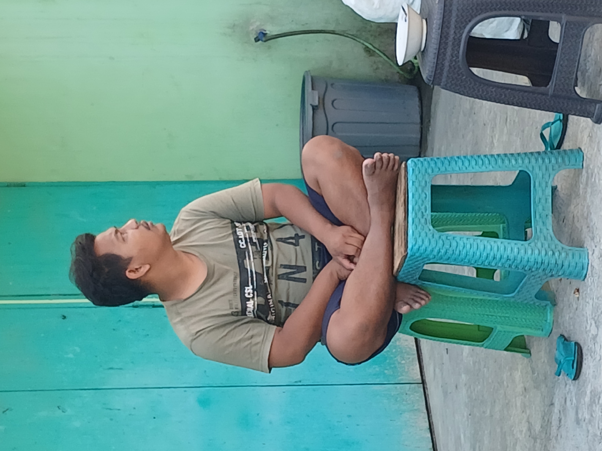 A man of Asian descent, appearing to be in his late 20s or early 30s, is seated cross-legged on a teal plastic stool. He wears a khaki t-shirt with a graphic print and dark blue shorts. His gaze is directed to the right, away from the camera. The man is positioned in front of a teal-colored wall or door. To the right of the man, there is a light green wall. A grey trash bin and a white bowl are visible against the green wall. A green hose is connected to a faucet on the wall. In the foreground, a pair of blue flip-flops lies on the concrete ground. A darker stool is also visible to the right of the teal stool. The scene appears to be outdoors, possibly in a residential area or a public space in Surakarta, Indonesia, given the architectural style and the environmental context.
