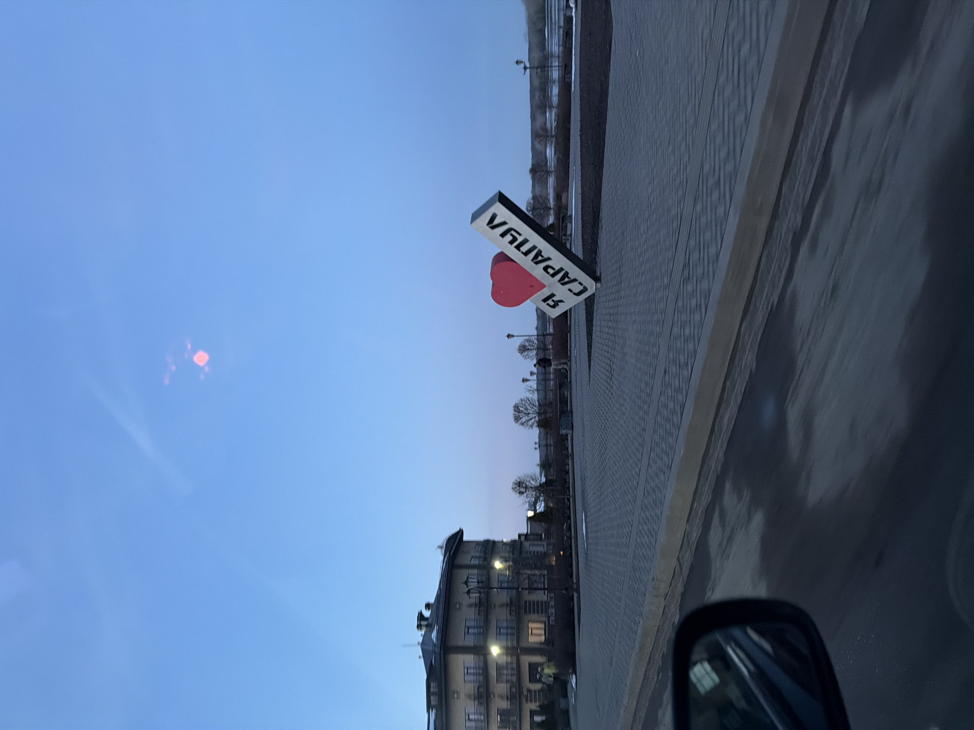 The image depicts a street scene in Sarapul, Russia. A prominent sign reading "Я люблю Сарапул" ("I love Sarapul") in Russian, with a red heart symbol, stands beside a brick-paved walkway. In the background, a multi-story building with lit windows is visible. Bare trees line the street beyond the building. The sky is a gradient of blue, suggesting twilight or dusk. The image is taken from inside a vehicle, as evidenced by the side-view mirror visible in the lower right corner. The overall scene suggests a public space within the city.