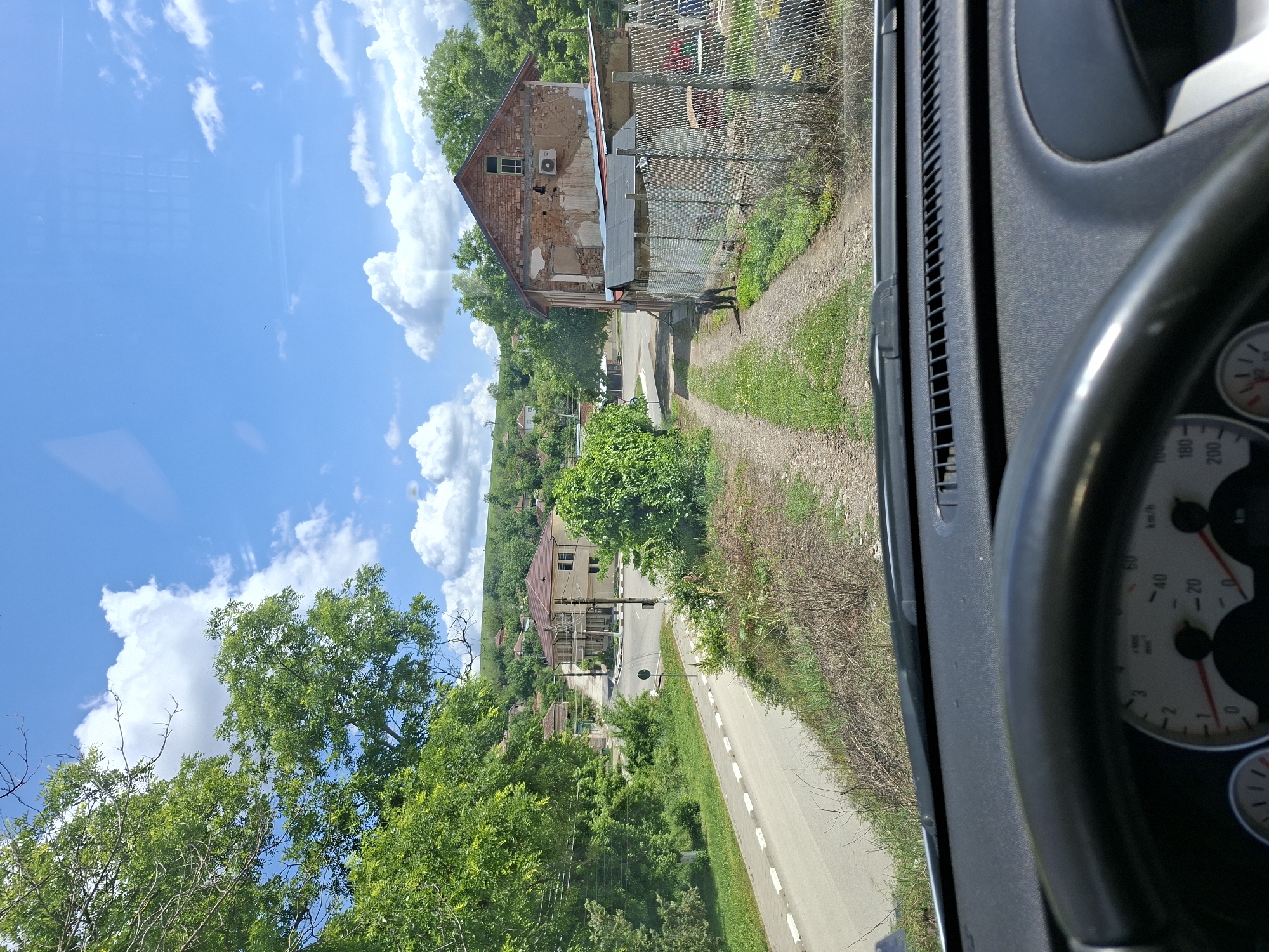 The image, taken from inside a vehicle, looks out onto a landscape in Bojuritsa, Bulgaria. The sky is blue with scattered white clouds. From left to right, a dense cluster of green trees occupies the lower portion of the image. A gray paved road with white edging runs parallel to the trees. Adjacent to the road, a low-lying patch of vegetation is visible. Further uphill, a light-colored building with a red-tiled roof is nestled amidst more trees and greenery.
In the top-right corner, a partially dilapidated brick building is visible behind a wire fence. A narrow, dirt path leads alongside the fence. A dark, elongated object, possibly an animal, is on the path. The interior of the vehicle is visible in the foreground. A portion of the dashboard and steering wheel obscure part of the view. The speedometer is partially visible, displaying kilometer per hour readings. The vehicle is not moving.