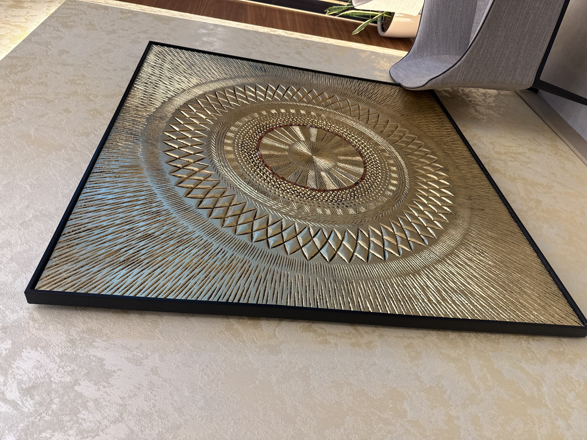 An intricately designed, square-framed artwork lies on a light-colored, textured floor, possibly a rug or carpet, in an indoor setting in Kazanlak, Bulgaria. The central focus is a large, golden, embossed or carved piece with a striking circular, mandala-like pattern.
The artwork features several concentric layers of design: a central, radially textured gold disc outlined by a thin, reddish-brown ring; a subsequent band of small, raised gold dots; a wider ring composed of raised, angular, diamond-like shapes; and an outermost section with numerous radial lines or grooves that extend to the black frame. The three-dimensional quality of the gold surface is enhanced by the ambient light, creating varied reflections and shadows that highlight the textures.
The black frame provides a sharp contrast to the luminous gold artwork. In the background, hints of a wooden surface, a green plant in a white pot, and a gray, fabric-covered item (possibly part of a chair or curtain) are visible. The lighting suggests daytime, with natural light illuminating the scene and casting soft shadows. There is no discernible activity taking place, nor is any text visible in the image.
