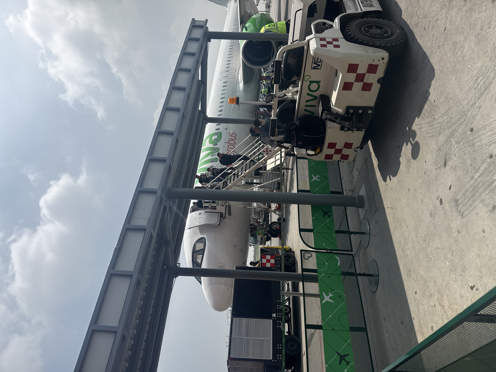 The image shows a Viva Aerobus airplane being boarded at an airport, likely in Apodaca, Mexico, based on the airline's presence there. Passengers are walking up the stairs to the aircraft. A baggage handling vehicle is parked nearby. The sky is overcast. The scene is viewed from within a boarding bridge structure. A ground services vehicle is visible with "Viva Aerobus" branding.