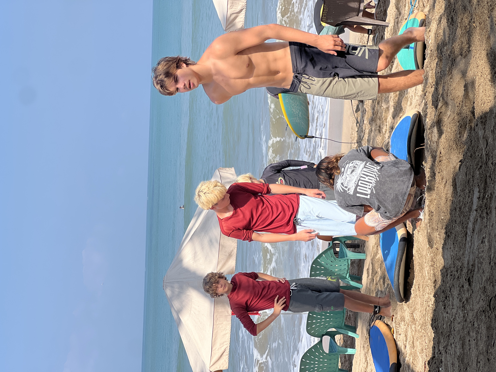 A group of young people, appearing to be in their late teens, are gathered on a sandy beach in Kuta, Indonesia, with surfboards and umbrellas visible. The ocean is to their left, with gentle waves and a clear blue sky above. The scene is bright and sunny, suggesting it is daytime.
In the foreground, a young person with light brown hair, wearing a dark grey t-shirt with "LOOKING FOR SUNSET" printed on the back, is kneeling and interacting with a blue surfboard. To their right, a young person with blonde hair, wearing dark swimming trunks, stands with their back to the camera, holding a yellow and blue surfboard.
Further left, two young people wearing red long-sleeved shirts are standing. The one on the left has curly brown hair and is wearing grey cargo shorts. The one on the right has blonde hair and is wearing light blue shorts. In the background, there are two large beige umbrellas, and several green plastic chairs are scattered on the sand. Another young person, shirtless and wearing dark swimming trunks, stands to the right, looking towards the ocean.
The beach appears to be a popular spot, with other individuals in the distance, some in the water. The overall atmosphere is relaxed and recreational.