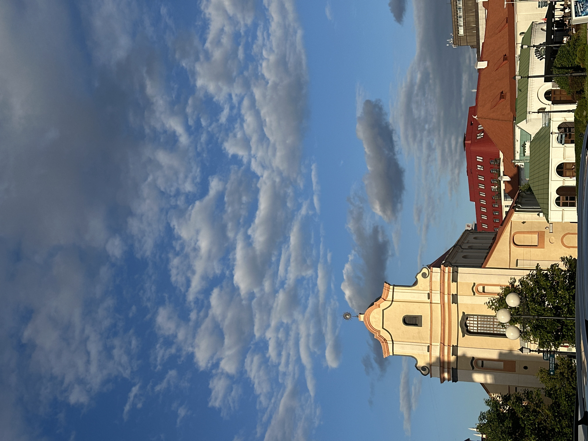 This image captures a view of the Church of St. Joseph in Minsk, Belarus, under a sky filled with scattered clouds. The church, a prominent baroque structure painted in light yellow and adorned with pink accents, is the focal point on the left side of the frame. Its distinctive facade features a stepped gable topped with a cross. To the right of the church, a cluster of buildings with orange and red roofs are visible, suggesting a dense urban setting. Several trees are interspersed among the buildings, and a modern street lamp with three spherical lights stands in the foreground. The scene is bathed in natural light, indicating it was taken during daylight hours, likely in the late afternoon or early evening, given the warm tones and the angle of the light. No people are clearly discernible in the image, and no specific actions or interactions are depicted. The overall impression is one of architectural beauty against a dynamic sky.