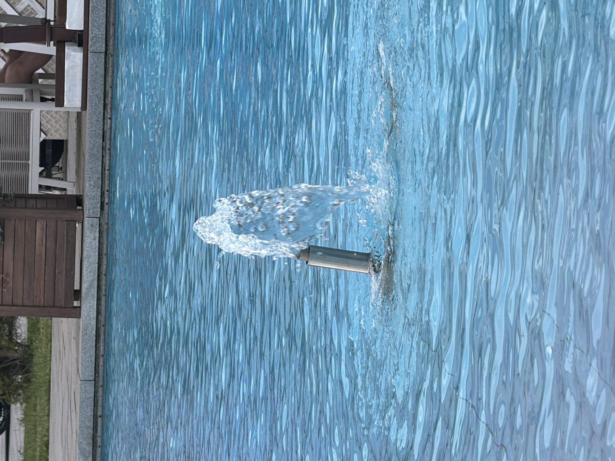 The image shows a close-up view of a fountain in a swimming pool. Water is spraying from a metal nozzle into the pool, creating ripples and splashes. The pool water is a vibrant blue color, and the surface is textured with wavy lines, suggesting movement. The nozzle of the fountain appears to be made of stainless steel.
In the background, to the left of the pool, there are glimpses of what appears to be a poolside area. We can see wooden planks, possibly part of a fence or deck, and some outdoor furniture, including a white chair. There is also a hint of greenery, suggesting an outdoor setting. The lighting suggests it is daytime, likely with sunlight reflecting off the water. There is no visible text in the image. The focus is on the dynamic action of the water and the texture of the pool.